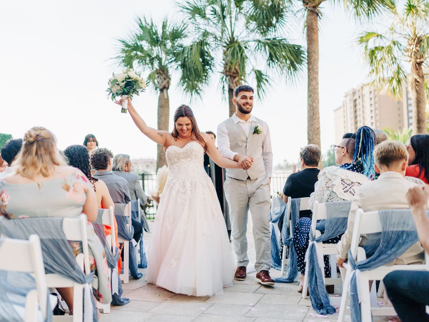 A wedding celebration on a palm-lined beachfront patio as newlyweds exit their ceremony with raised arms amidst seated guests.
