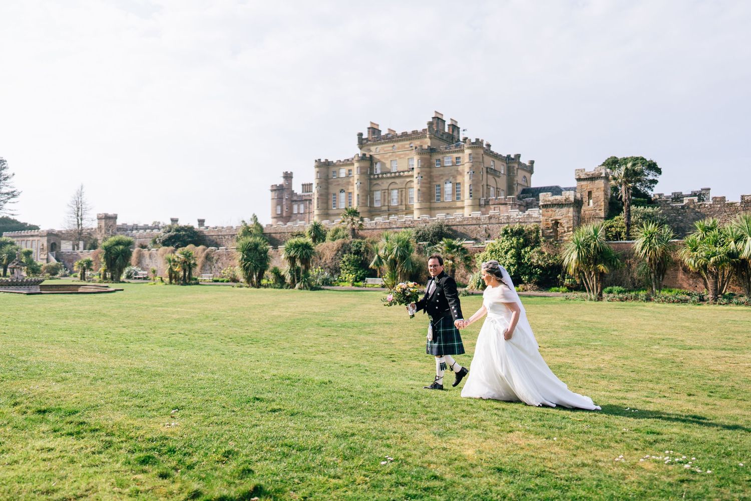 Two people in wedding attire walk across a green lawn with a majestic stone castle and gardens in the background.