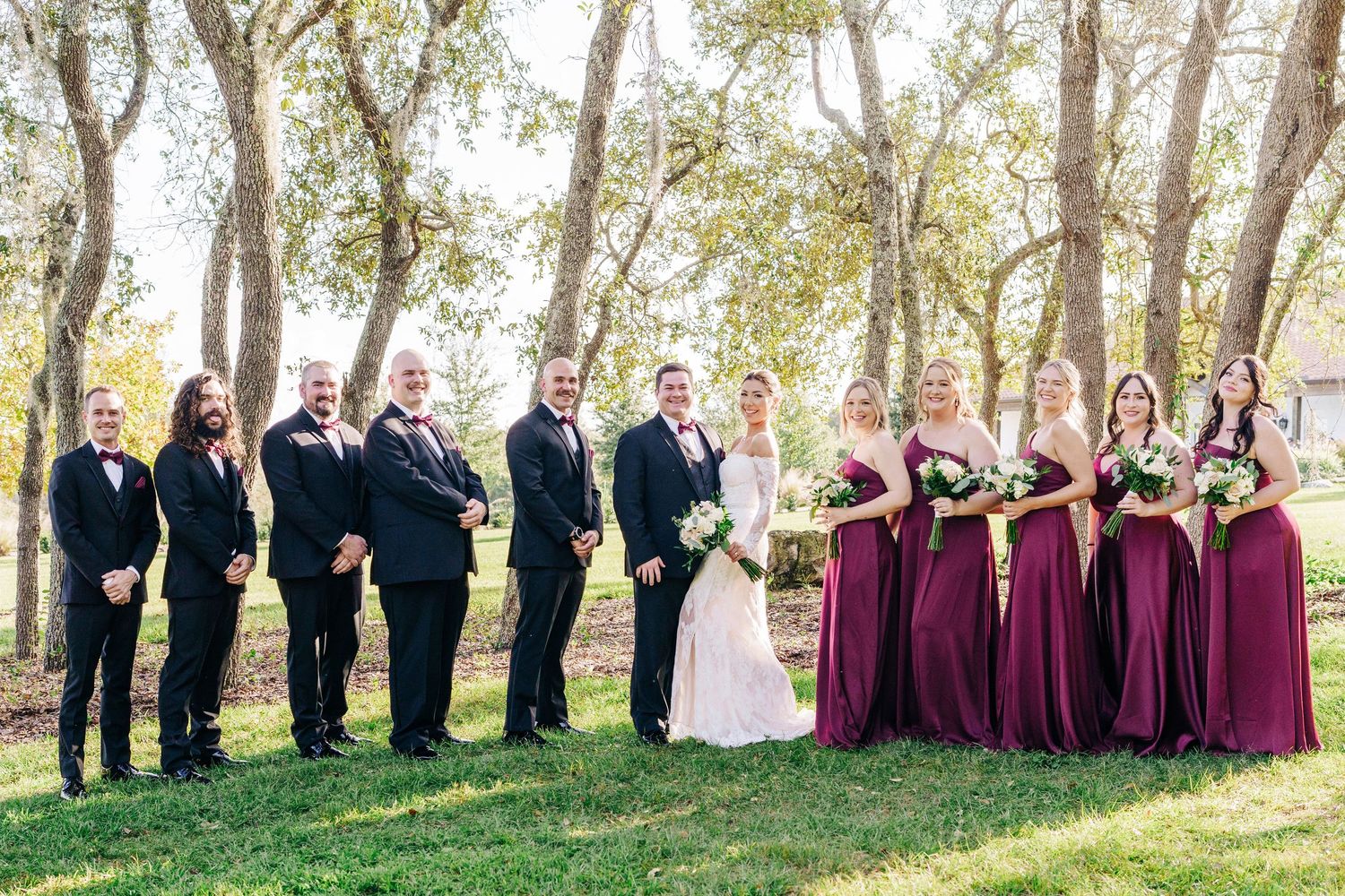 Wedding party in formal attire poses outdoors with bridesmaids in burgundy dresses and groomsmen in dark suits.
