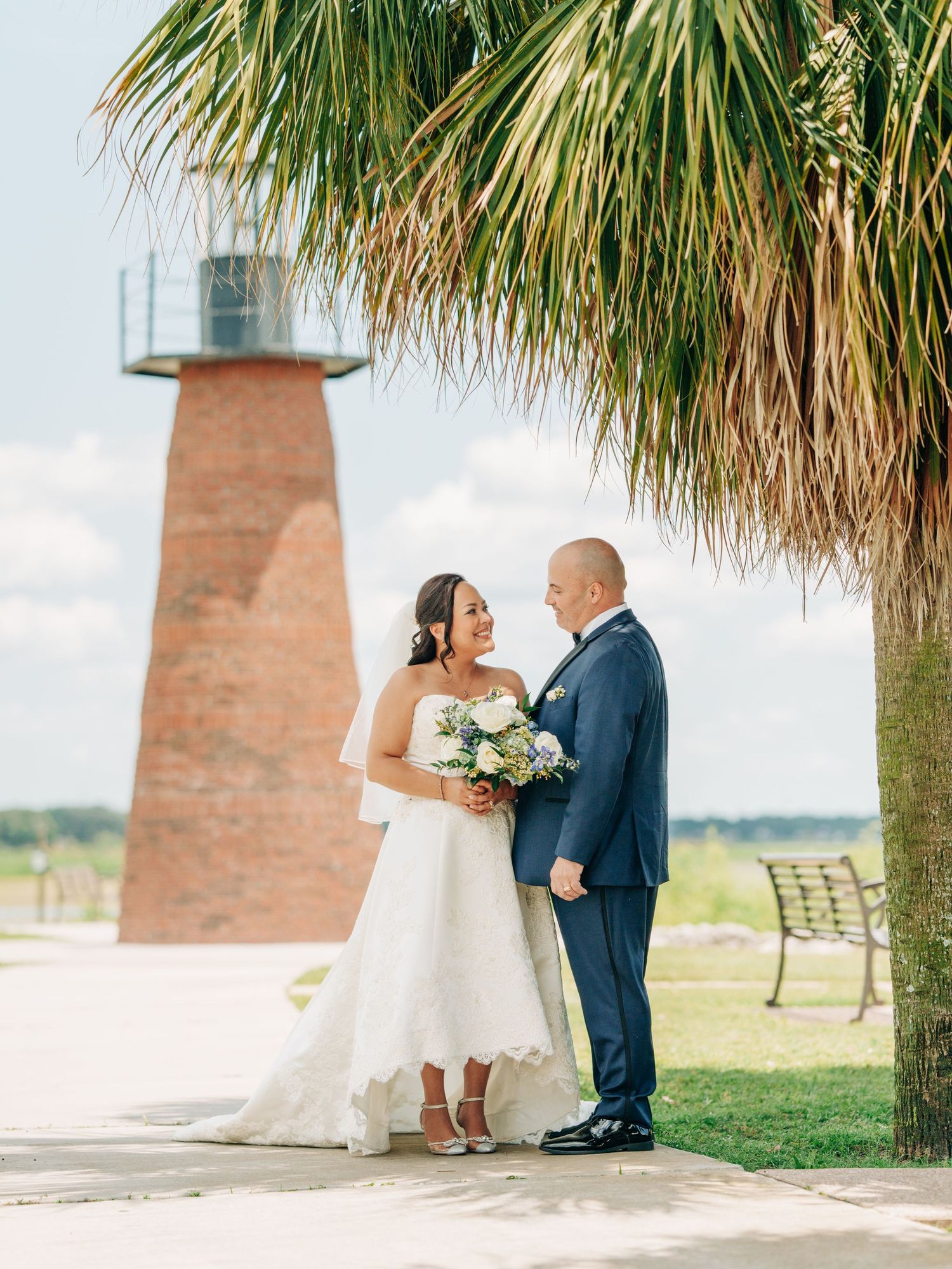 A romantic wedding moment at a lighthouse with palm trees and a bench in the background.