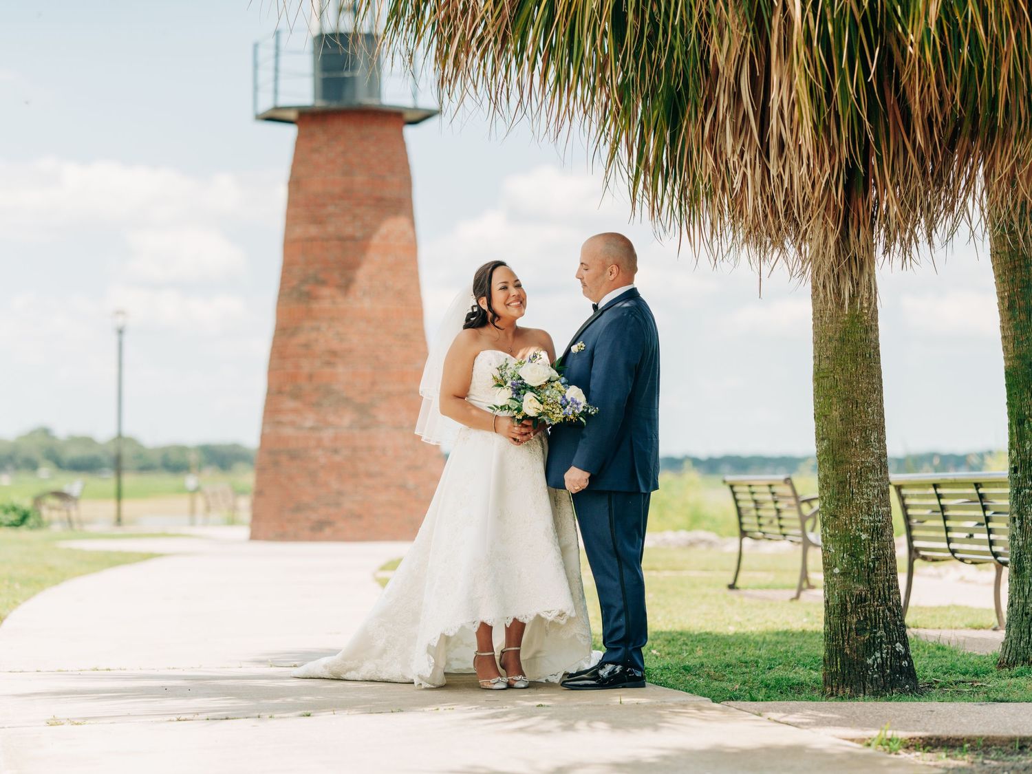 A couple in a navy suit and white wedding dress stand near a brick lighthouse and palm tree on their wedding day.