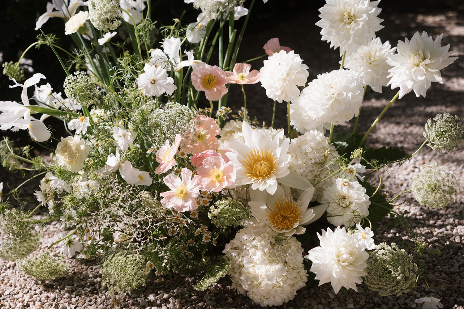Beautiful white dahlias and delicate pink flowers arranged with soft baby's breath on a gravel surface in natural light.