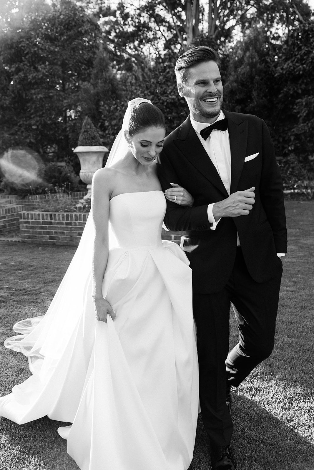 Black and white wedding photo of a bride in strapless gown with flowing train walking arm in arm with groom in tuxedo outdoors.