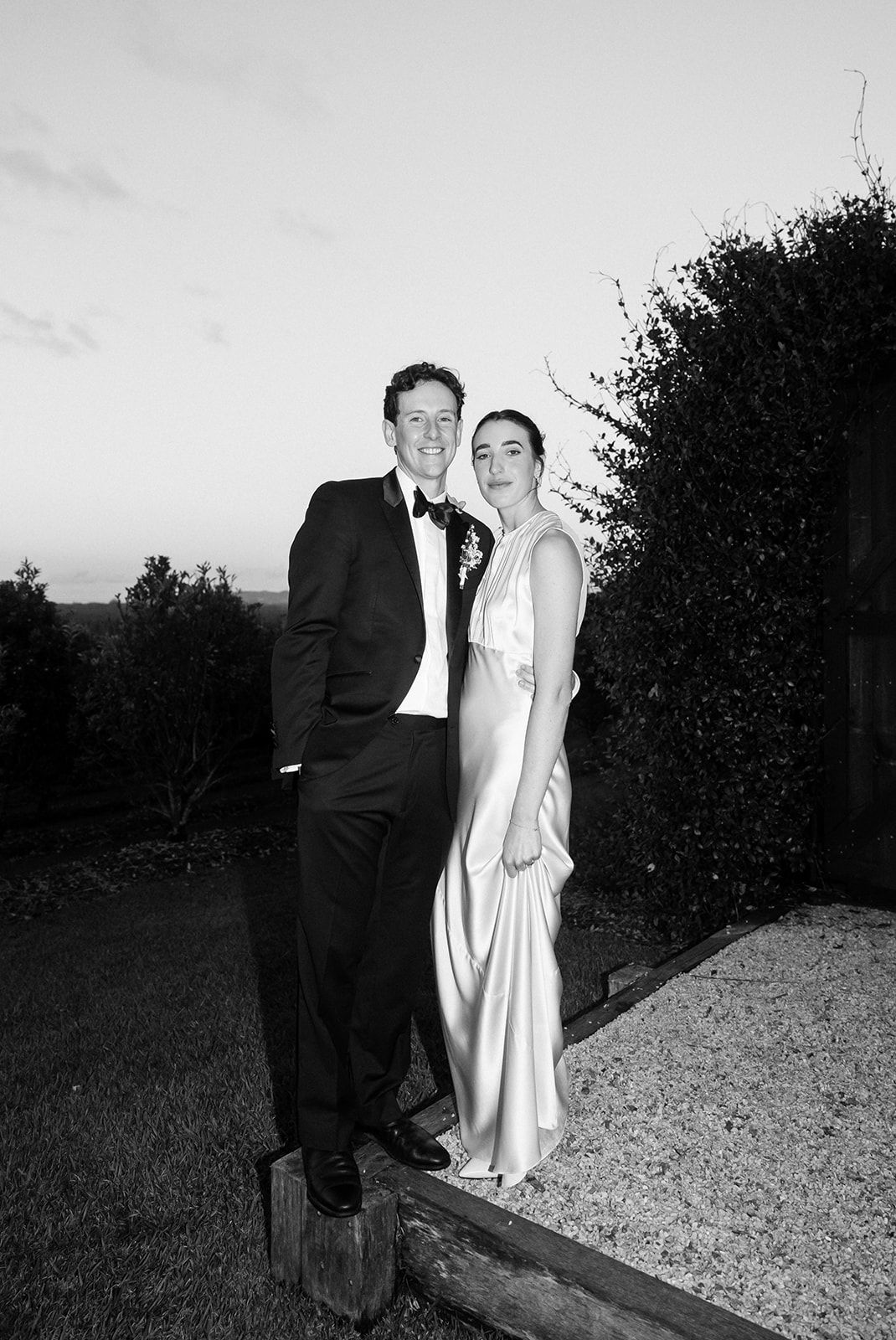 A couple in formal evening wear stands on a wooden fence at dusk, captured in black and white photography.