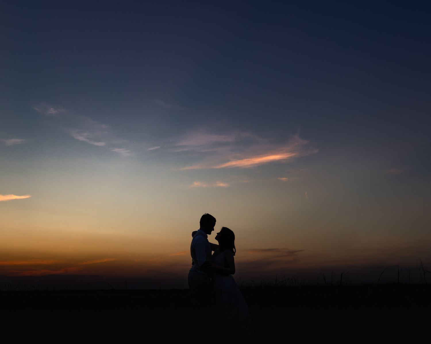 Deux silhouettes s'embrassent dans un ciel de coucher de soleil spectaculaire avec des teintes orange et violettes à l'horizon.