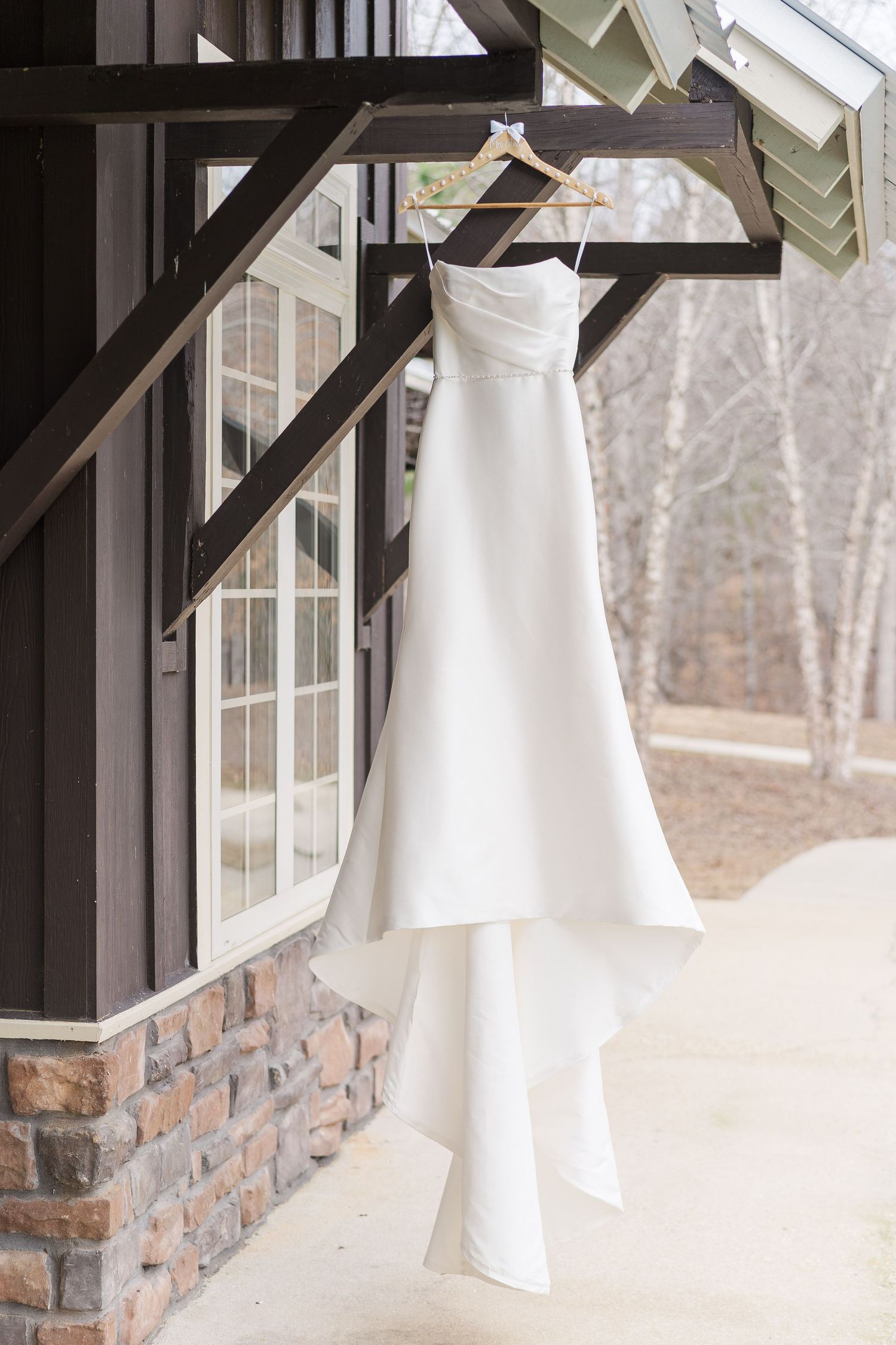 A white strapless wedding dress hangs elegantly on a wooden beam against stone exterior and windows of a rustic building.