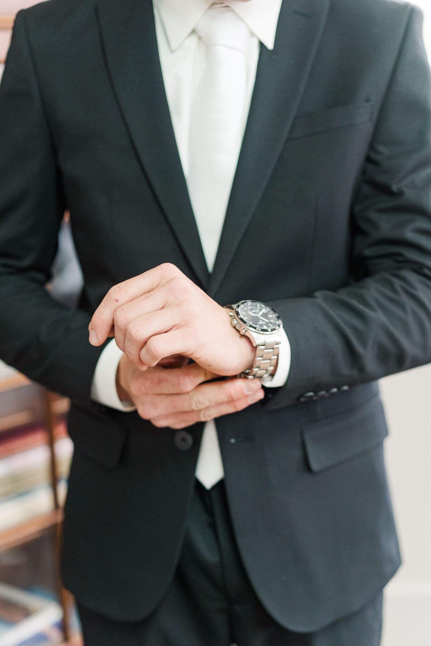 A person wearing a black suit and white tie checks their silver watch with both hands.