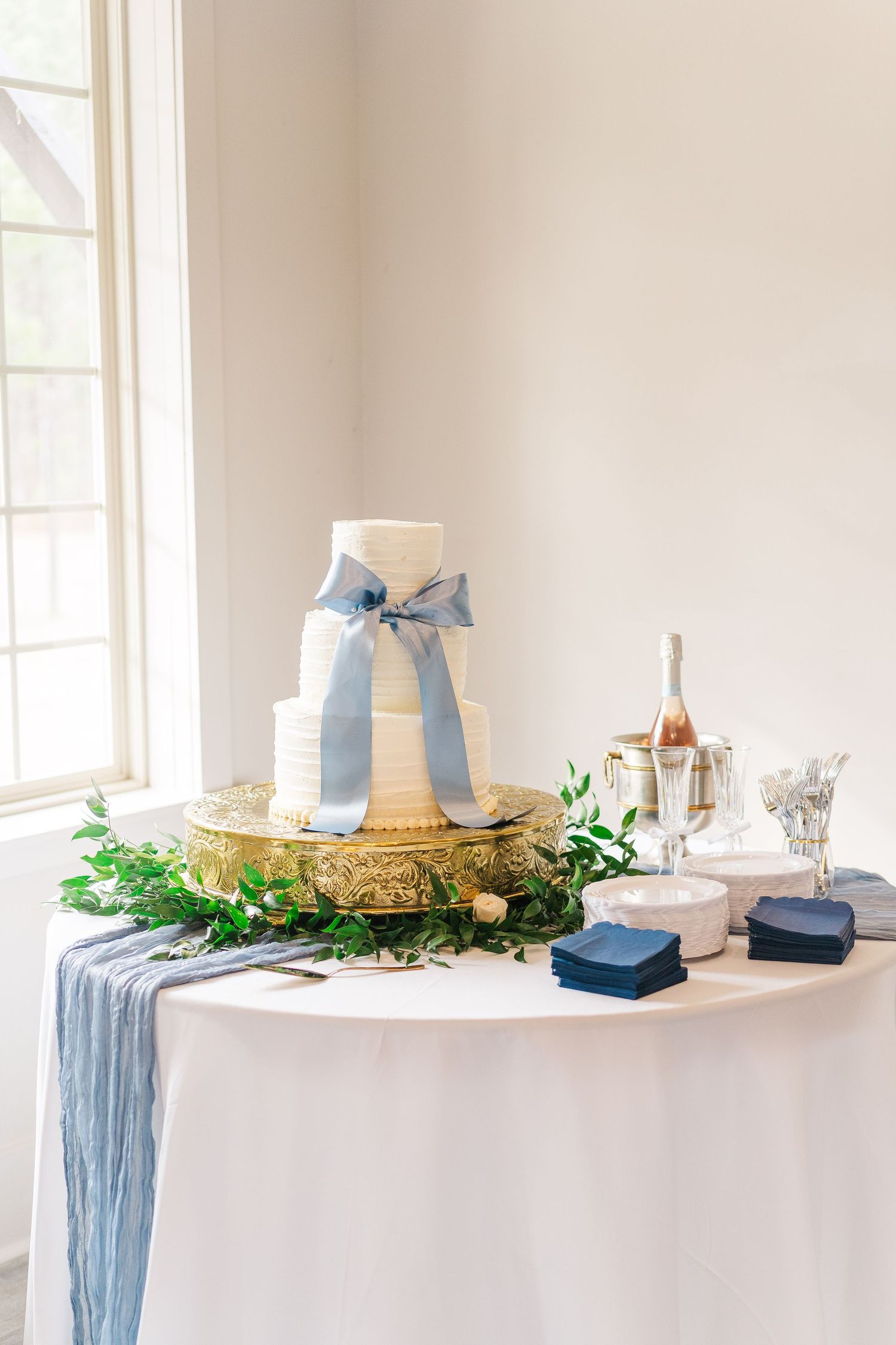 Elegant white wedding cake with light blue silk ribbon on ornate gold stand with greenery and champagne table setting.