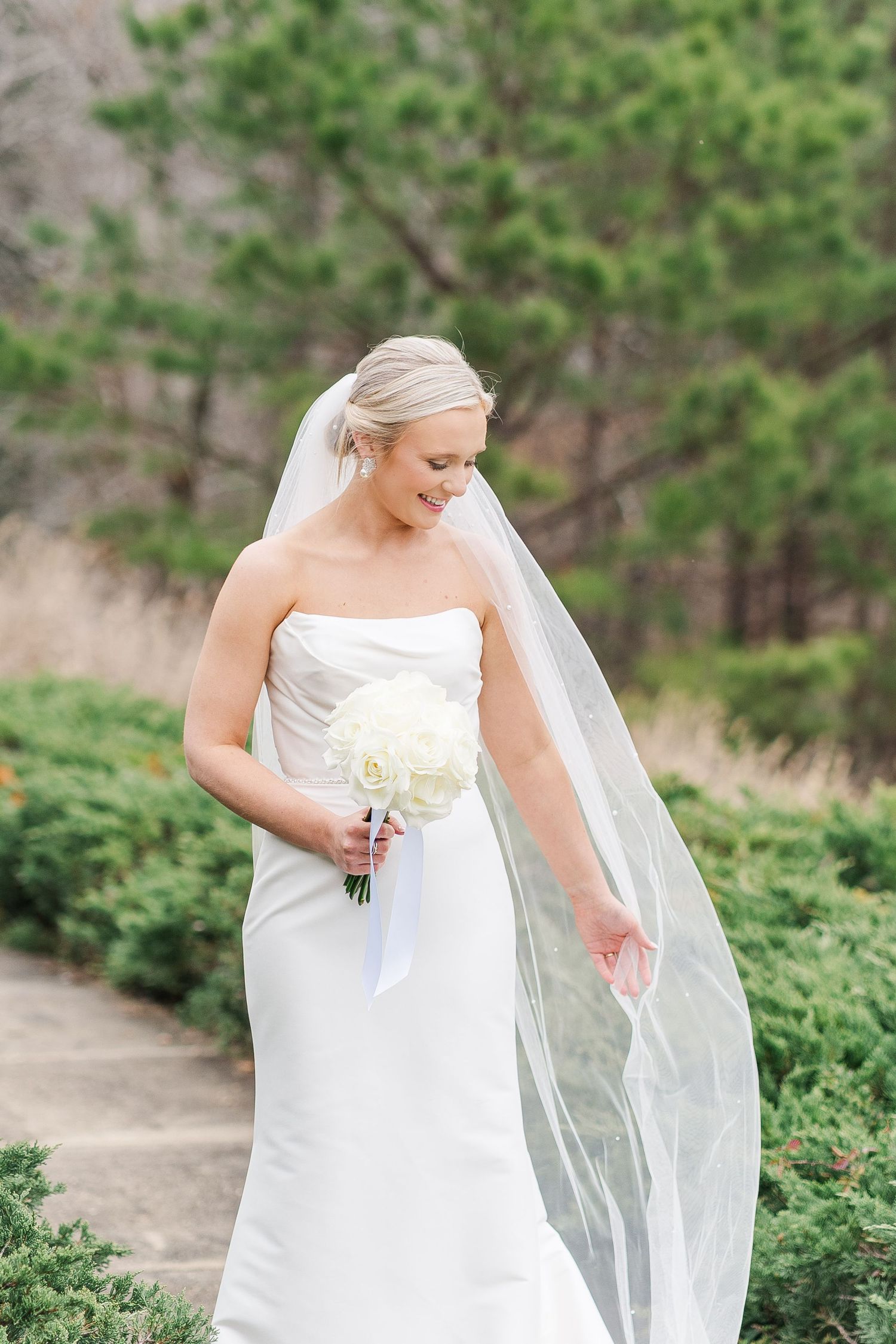 Beautiful bride in white strapless gown holds white bouquet while wearing flowing cathedral veil in outdoor garden setting.