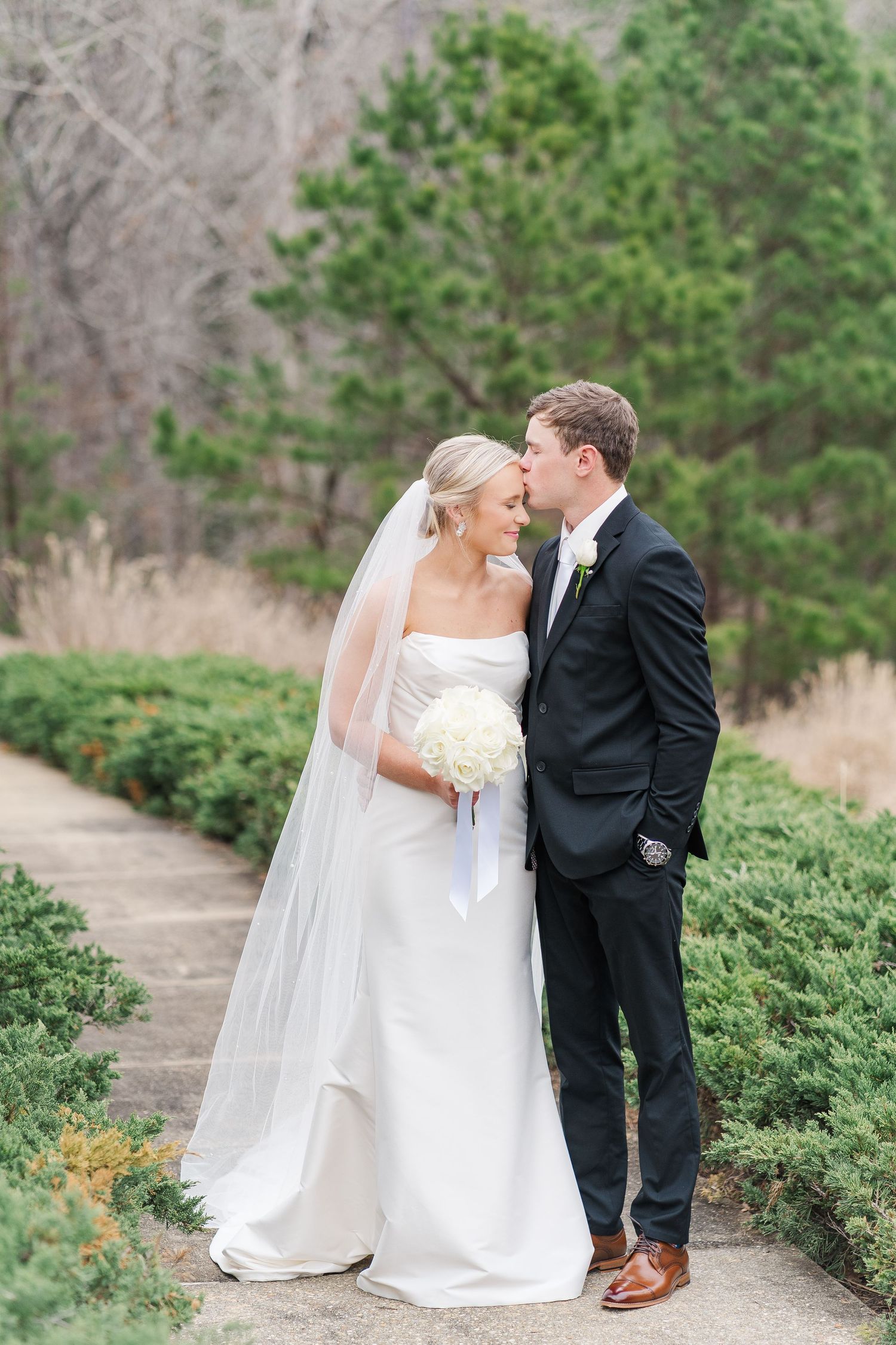 Newlywed couple shares intimate moment on stone path surrounded by green shrubbery and evergreen trees.
