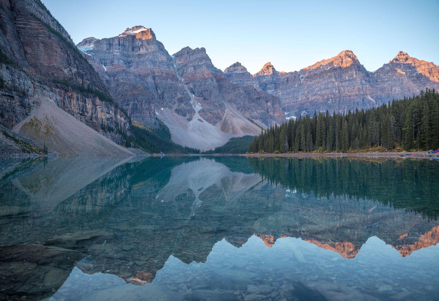 Mirror-like reflection of mountain peaks in calm waters of Moraine Lake during sunrise.