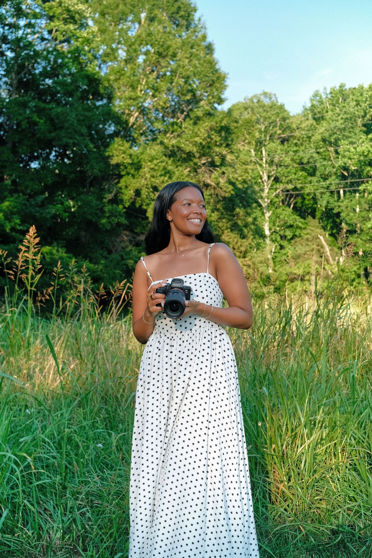 A photographer in a white polka dot maxi dress smiles while holding a camera in a sunny meadow with trees in background.