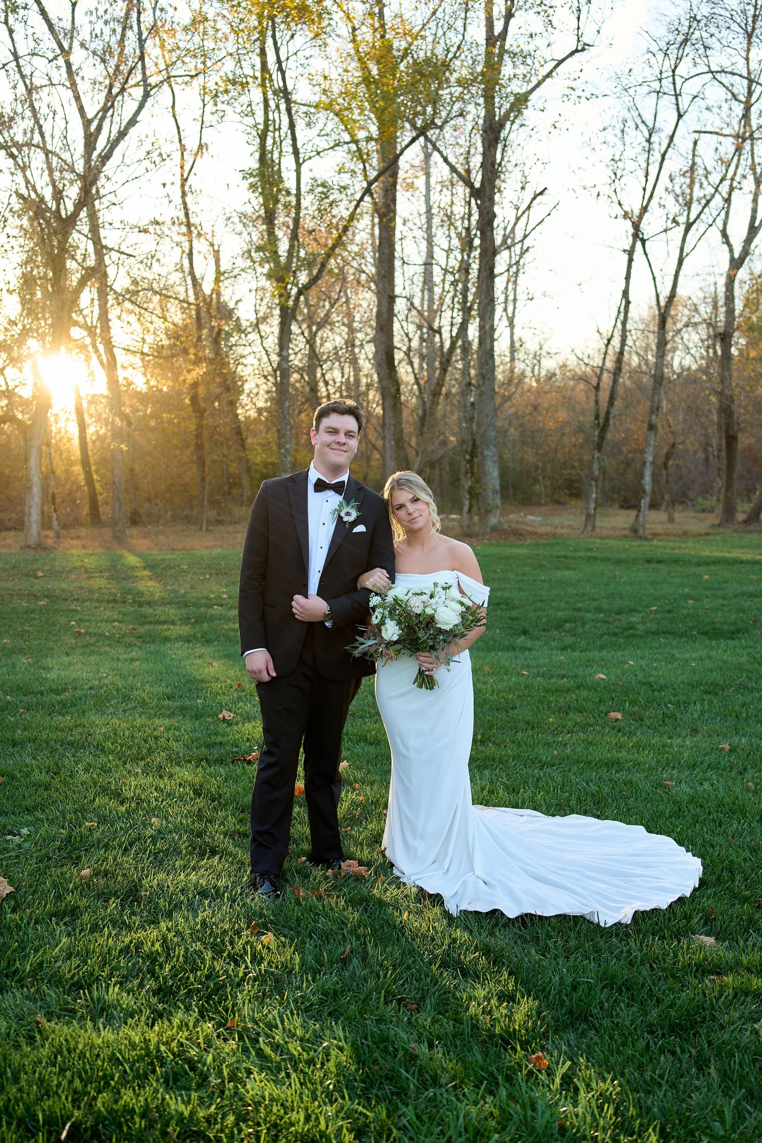A bride and groom pose together on a grass lawn at sunset with trees and golden light in the background.