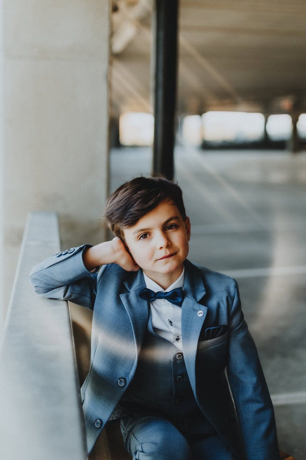 Een jonge jongen in een blauw pak met een strikje kijkt vrolijk in de camera voor zijn communie foto's in een parking bij Torhout