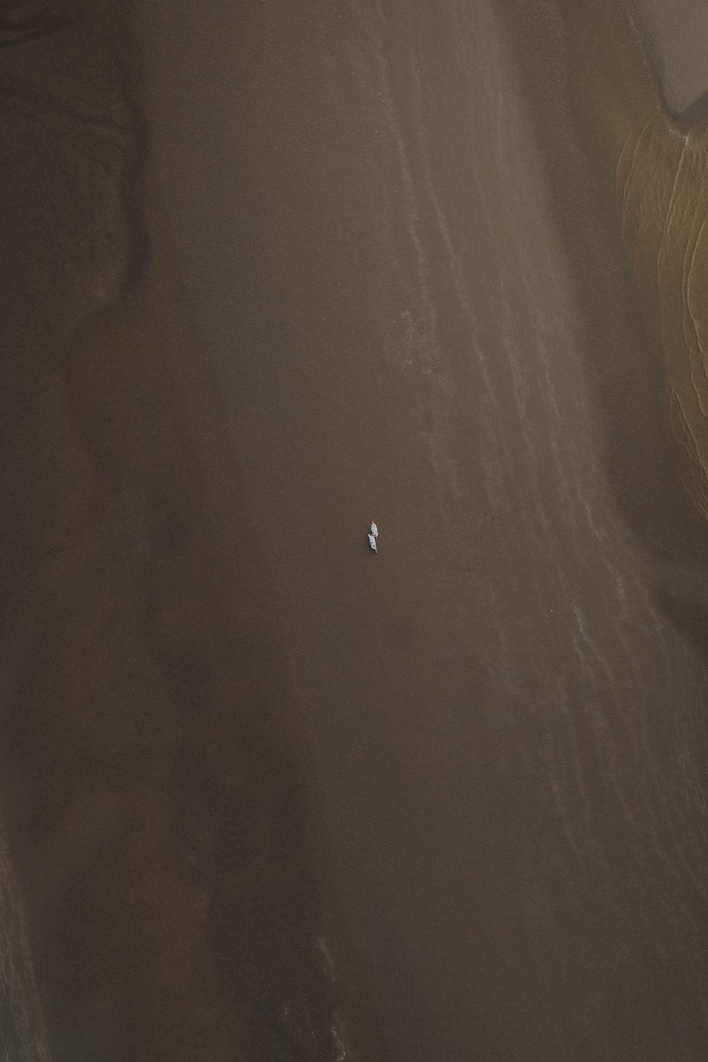 Luchtfoto van een koppel liggend op een donker zandstrand. Het zand toont zachte patronen van water en wind. Rustig en intiem moment.
