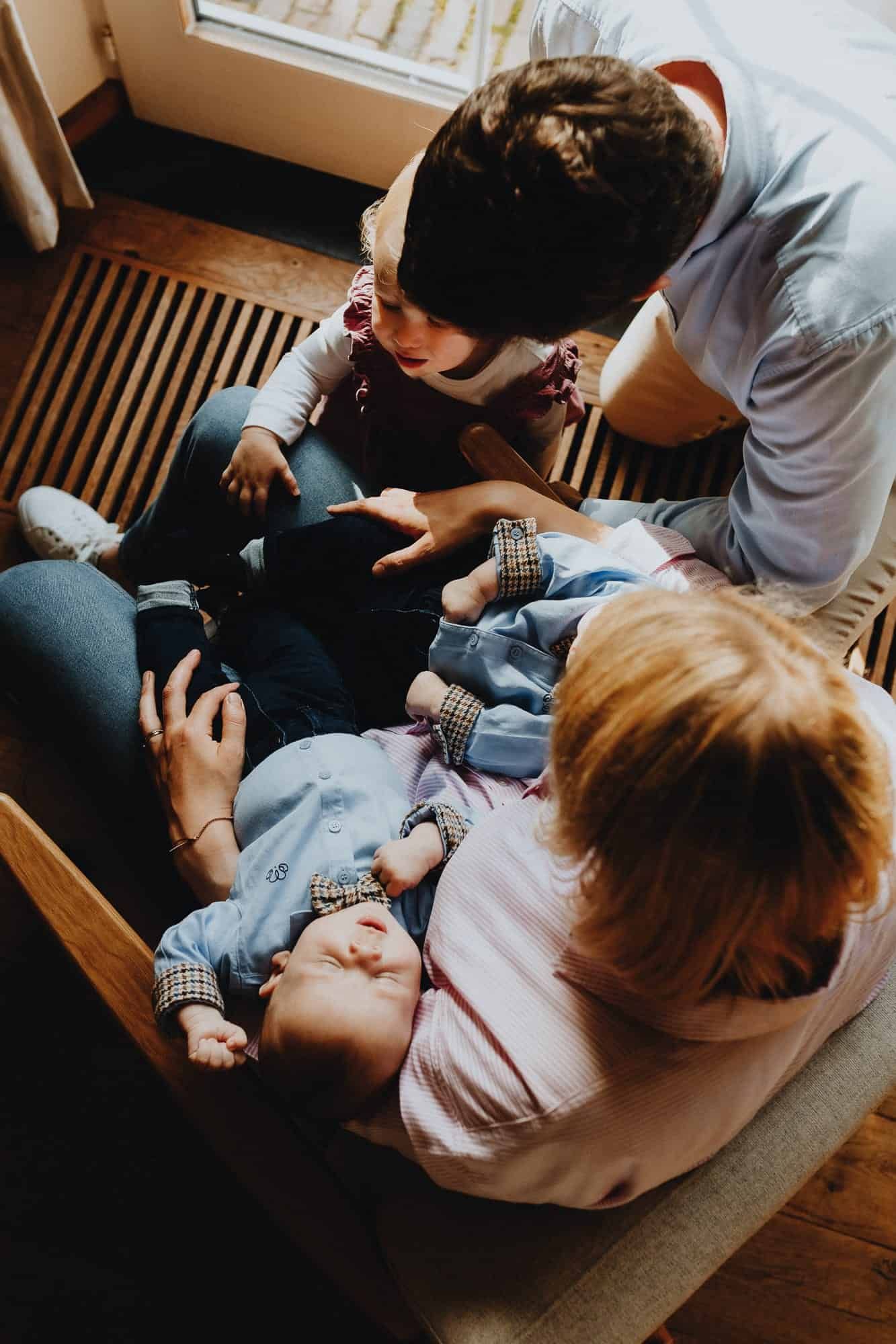 Een gezin verzamelt zich rond een stoel. Een volwassene houdt een slapende baby vast terwijl een peuter toekijkt. Warm en teder moment.