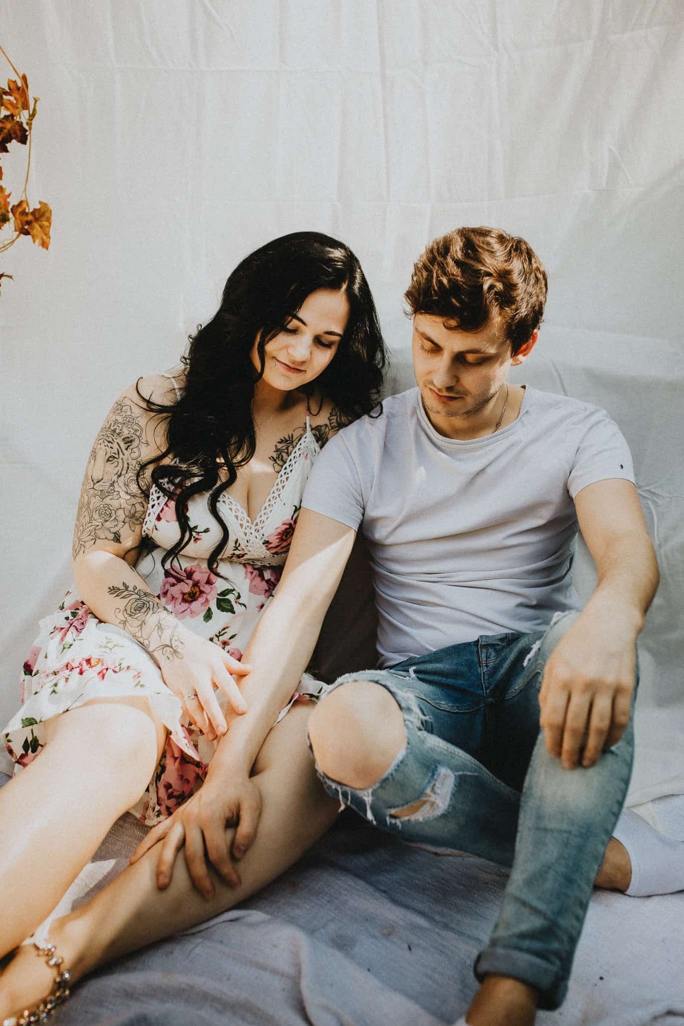 Een vrouw in bloemenjurk en een man in wit hemd en gescheurde jeans zitten dicht op een lichte stoffen achtergrond. Serene, intieme sfeer.