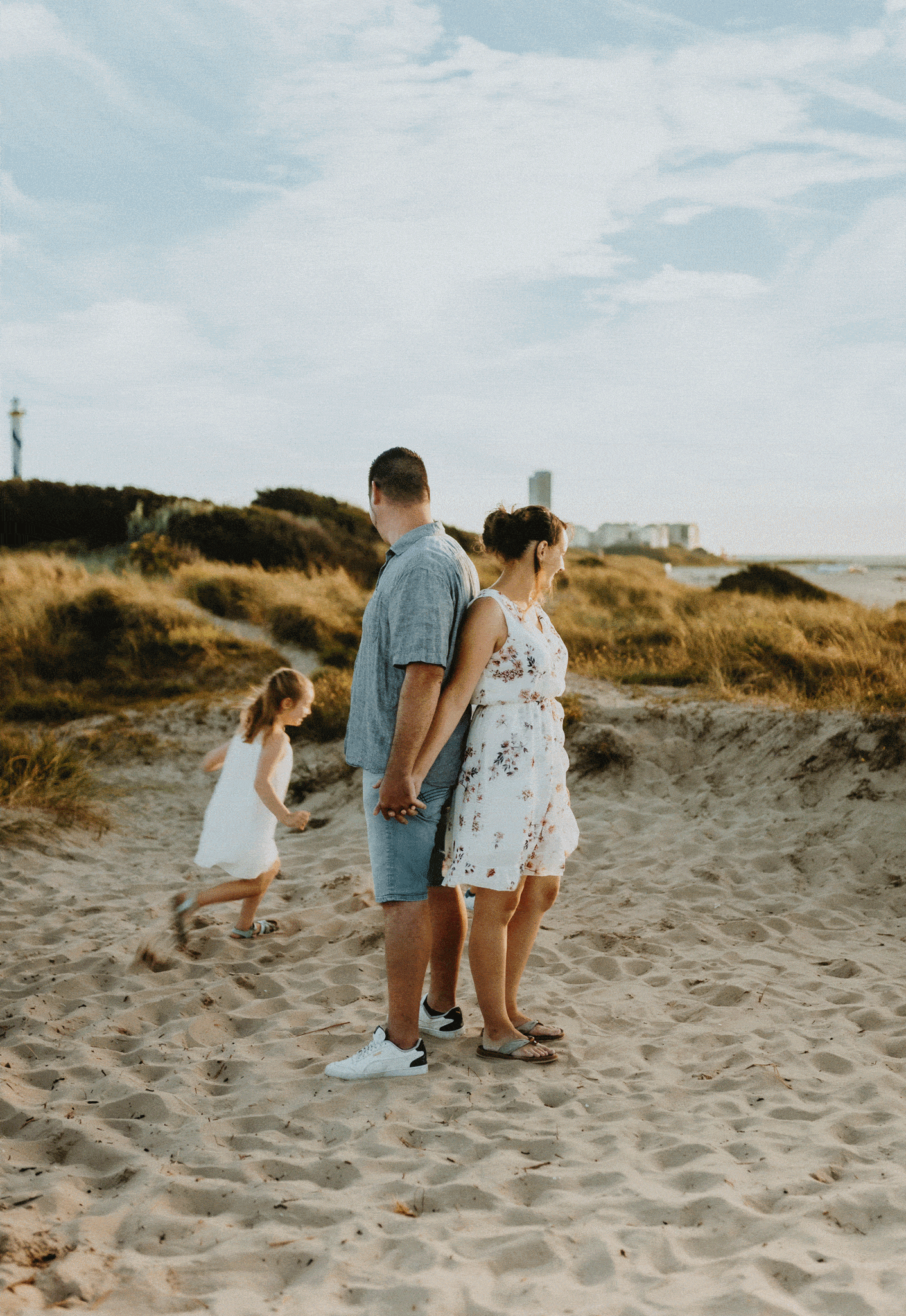 Een koppel houdt elkaars hand vast op het strand. Kind in witte jurk rent langs grasduinen en een vuurtoren in de verte.
