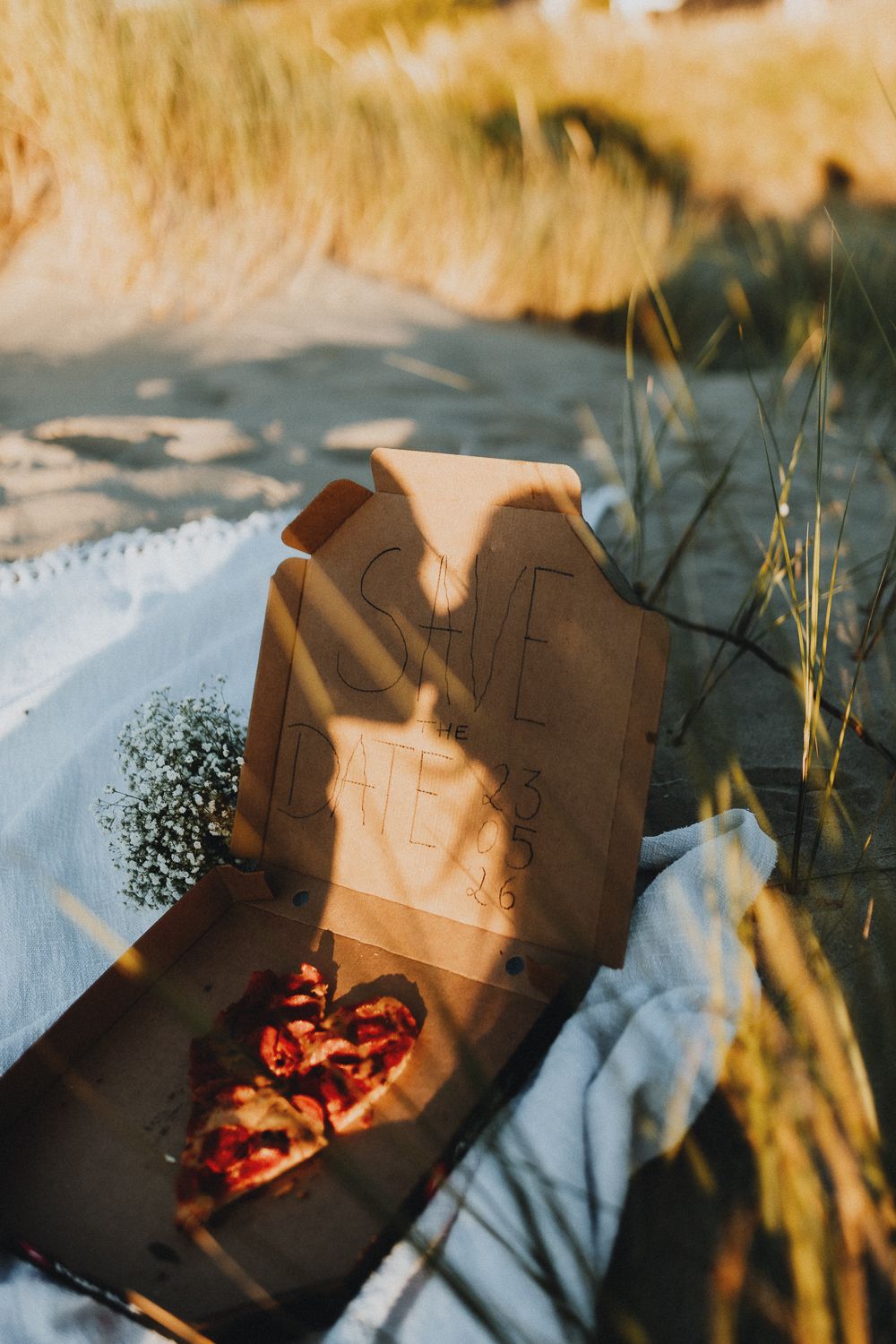 Open pizzadoos op het strand met ‘Save the Date 23-05-26’ erin geschreven. Hartvormige pizza en gipskruid naast de doos.