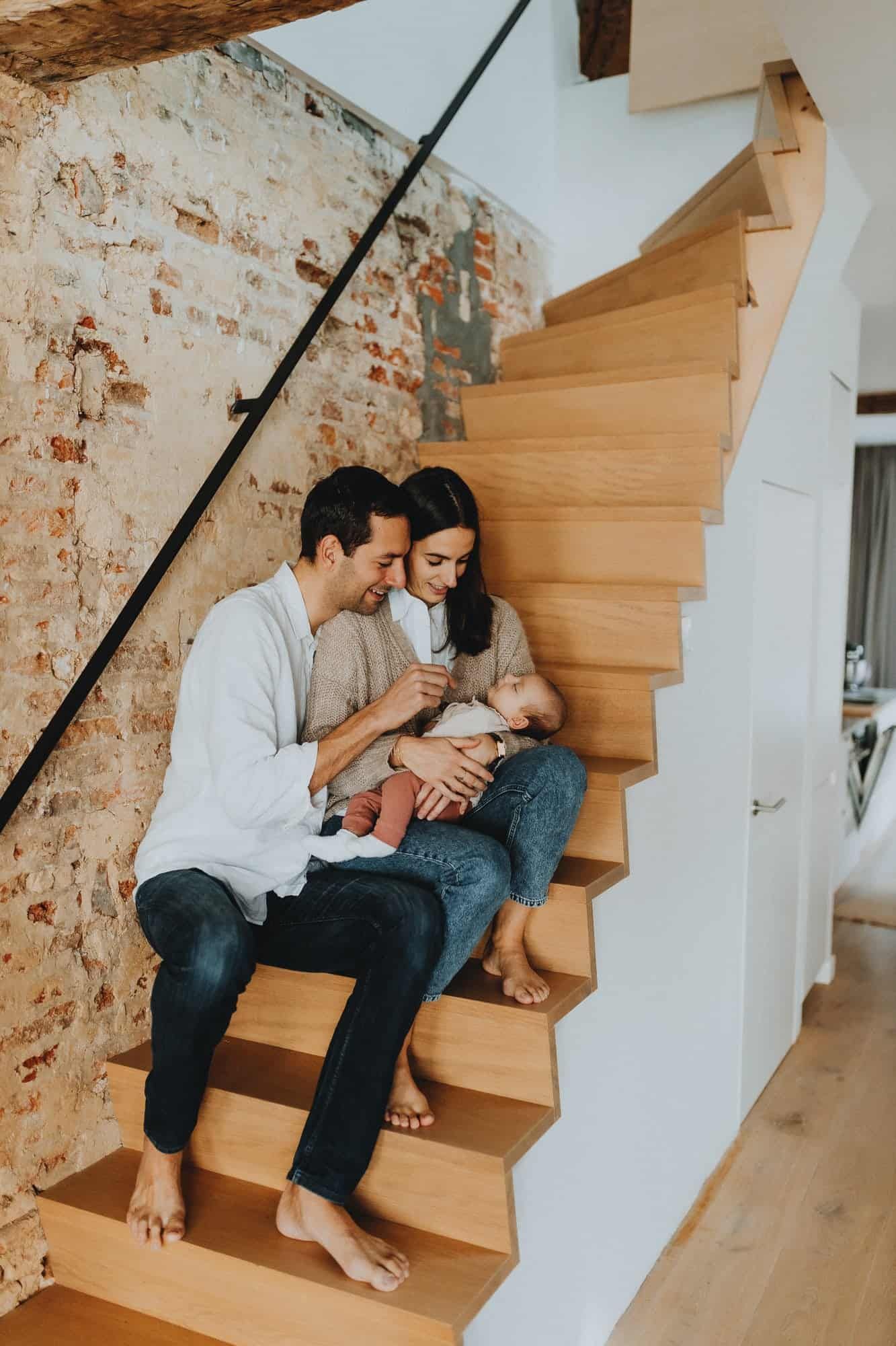 Een koppel zit op houten trappen in Gent, voor een bakstenen muur, en kijkt liefdevol naar hun baby. Warm en intiem gezinsmoment.