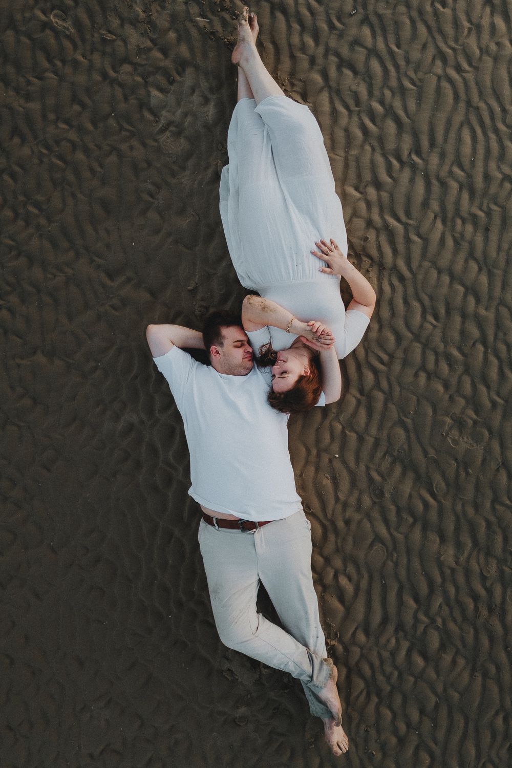 Een koppel in witte kleding ligt dicht tegen elkaar op het zand. Het gestructureerde zand en hun ontspannen blik creëren een intiem moment.