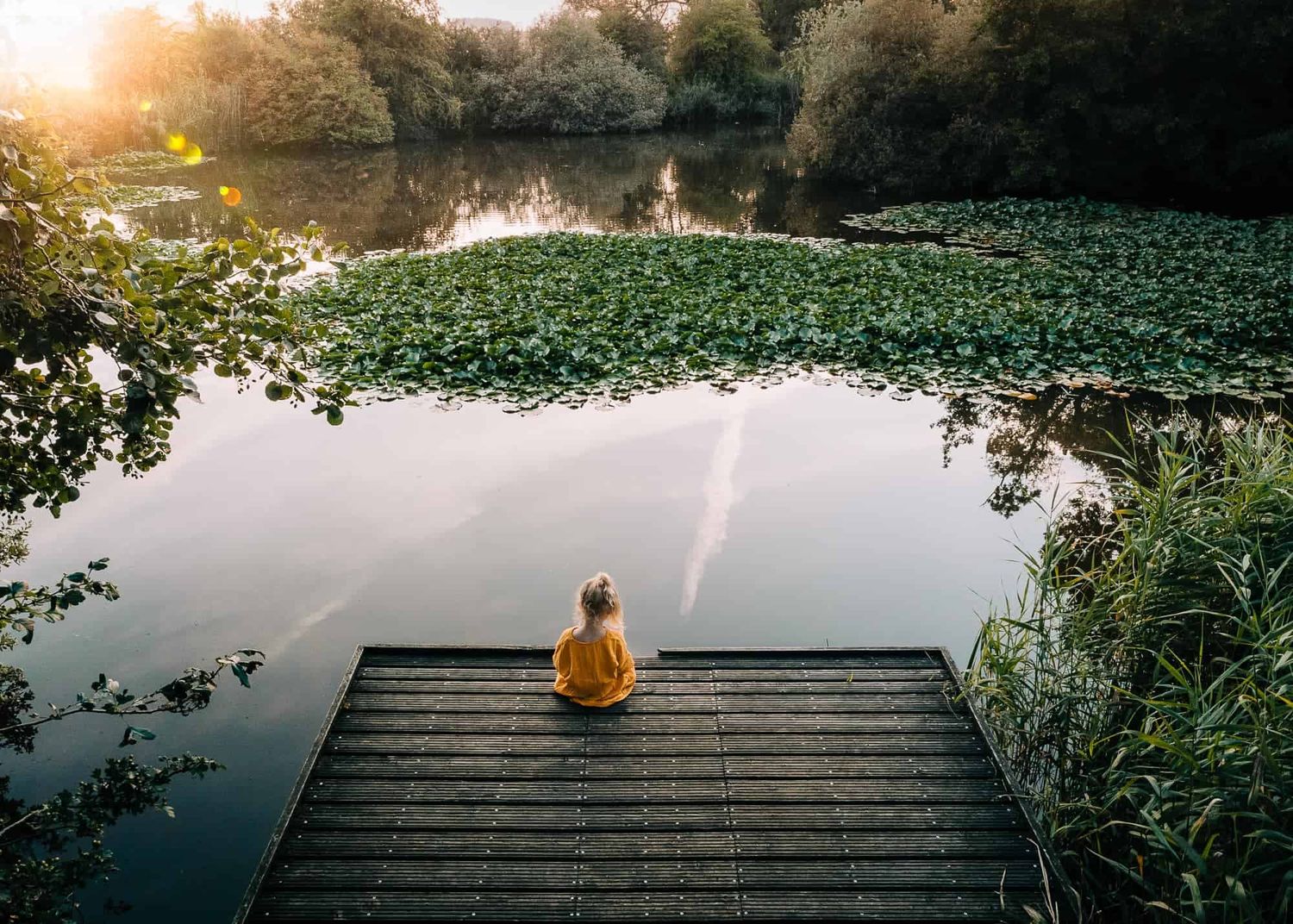Een klein meisje in een gele jurk zit op een houten steiger aan een vijver met lelies, in een groen gebied nabij Diksmuide bij zonsondergang