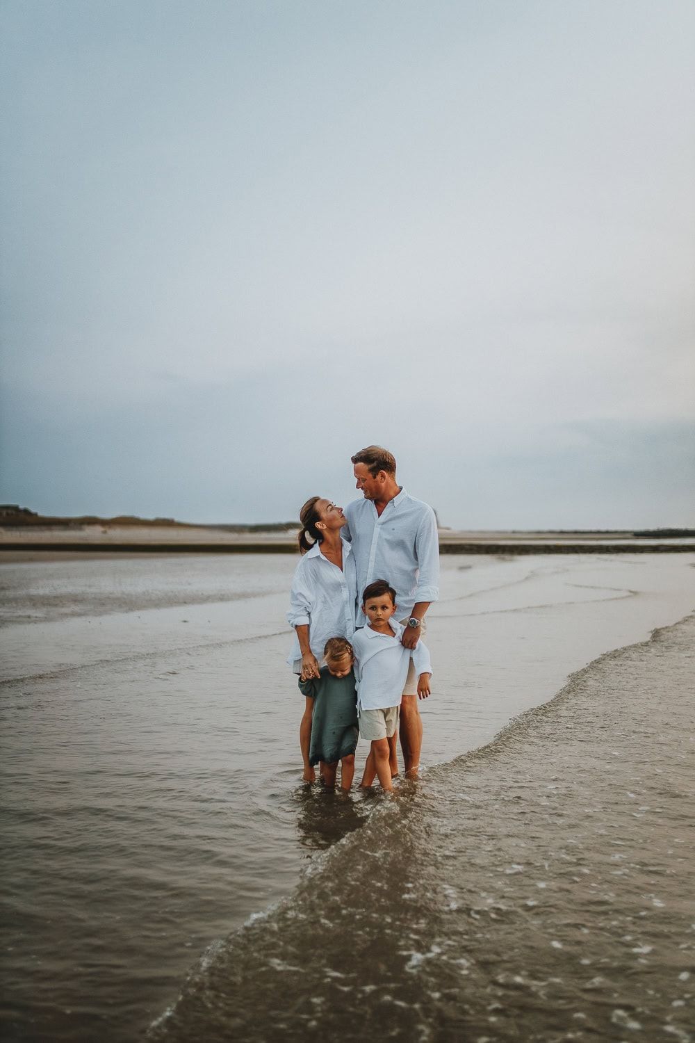 Een gezin van vier staat op het strand, lachend naar elkaar. Ouders dragen witte hemden, kinderen staan tussen hen in. Rustige sfeer.
