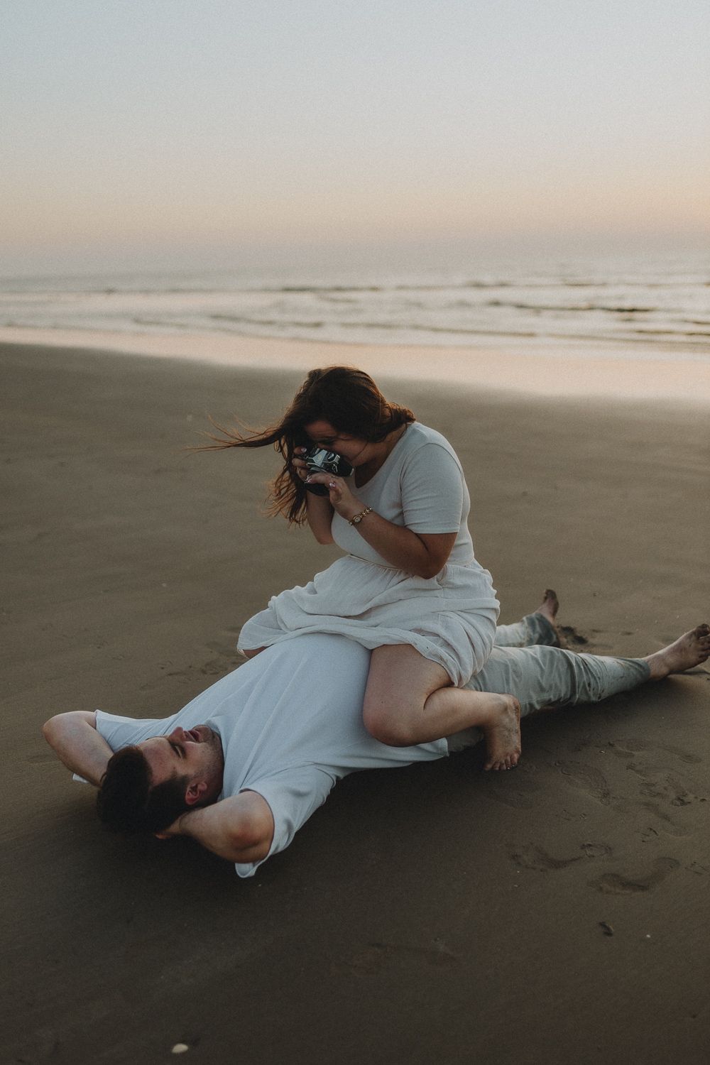 Vrouw zit lachend op haar partner en maakt een foto met een vintage camera – spontane koppelfoto aan zee in Oostende, België.