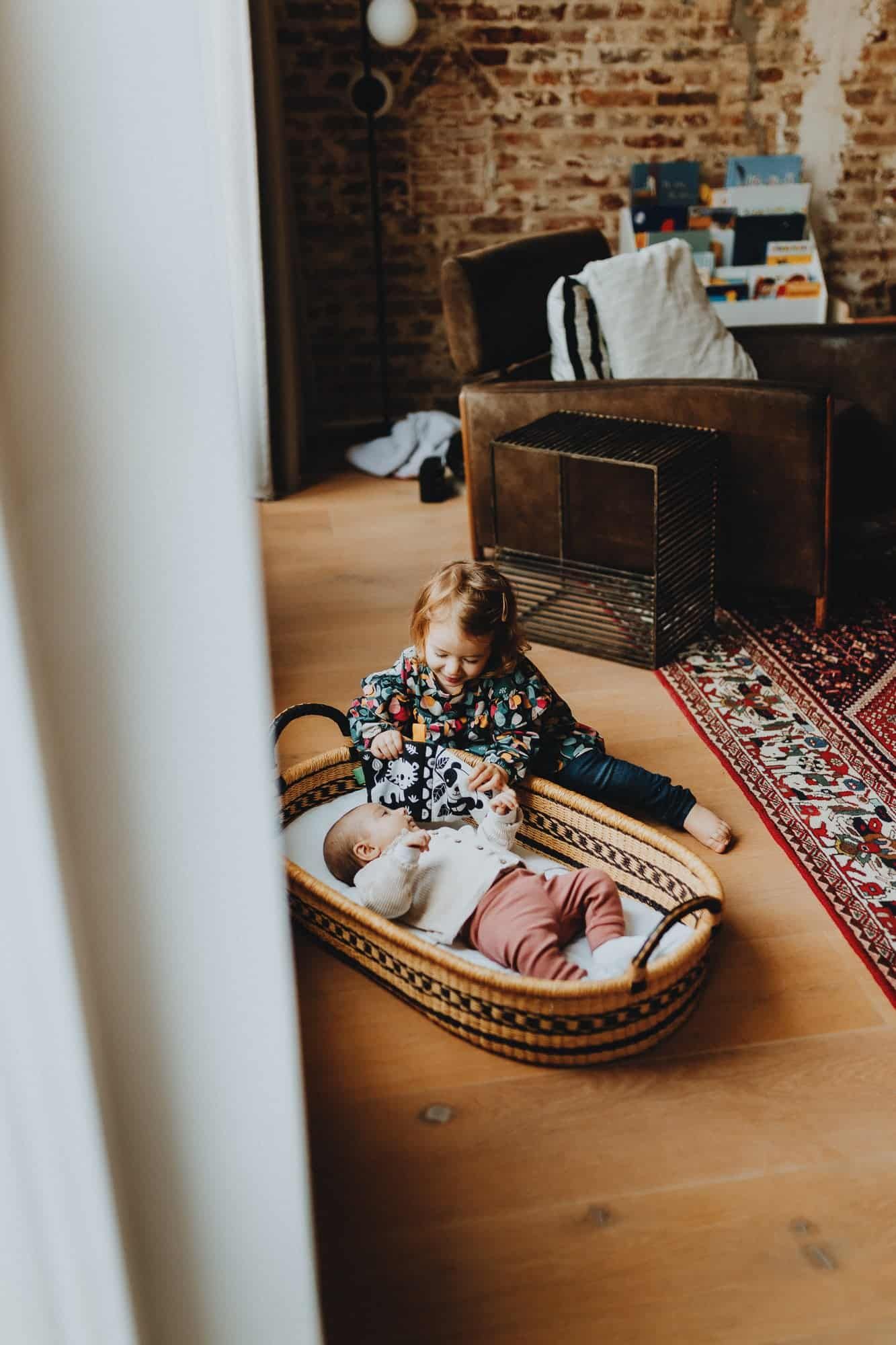 Gezinsmoment in een woning in Ieper: peuter speelt op houten vloer met een baby in een mand. Warme sfeer en huiselijke setting.