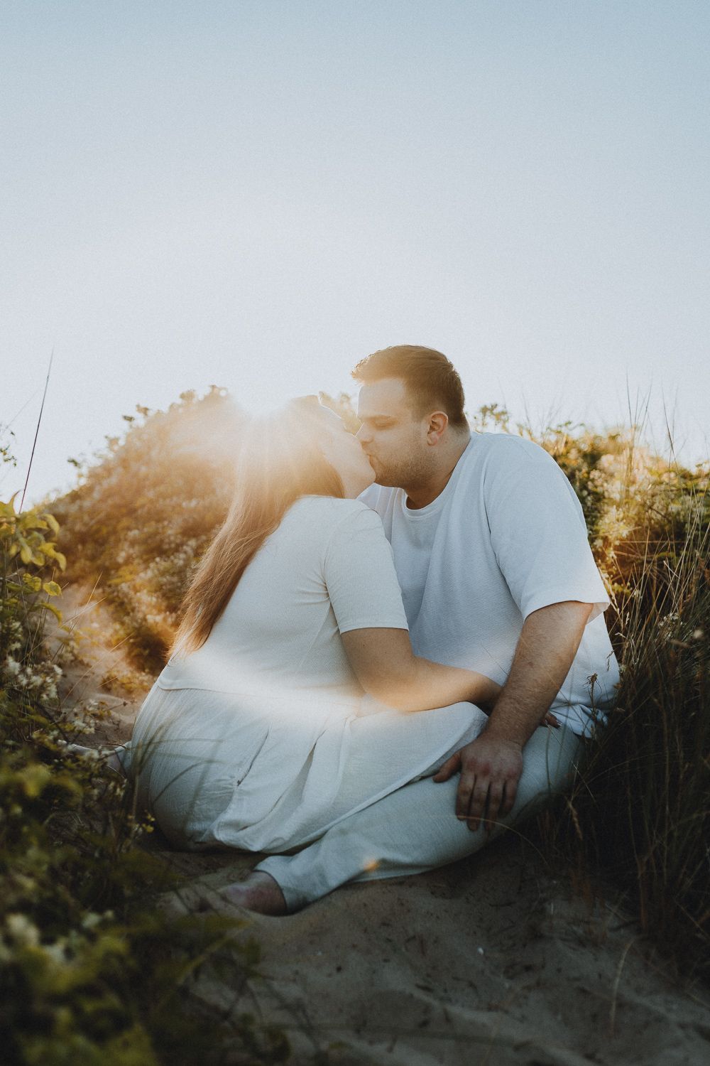 Een koppel in witte kleren deelt een tedere kus op een zandduin, badend in warm zonlicht. Liefdevol en sereen moment vol rust.