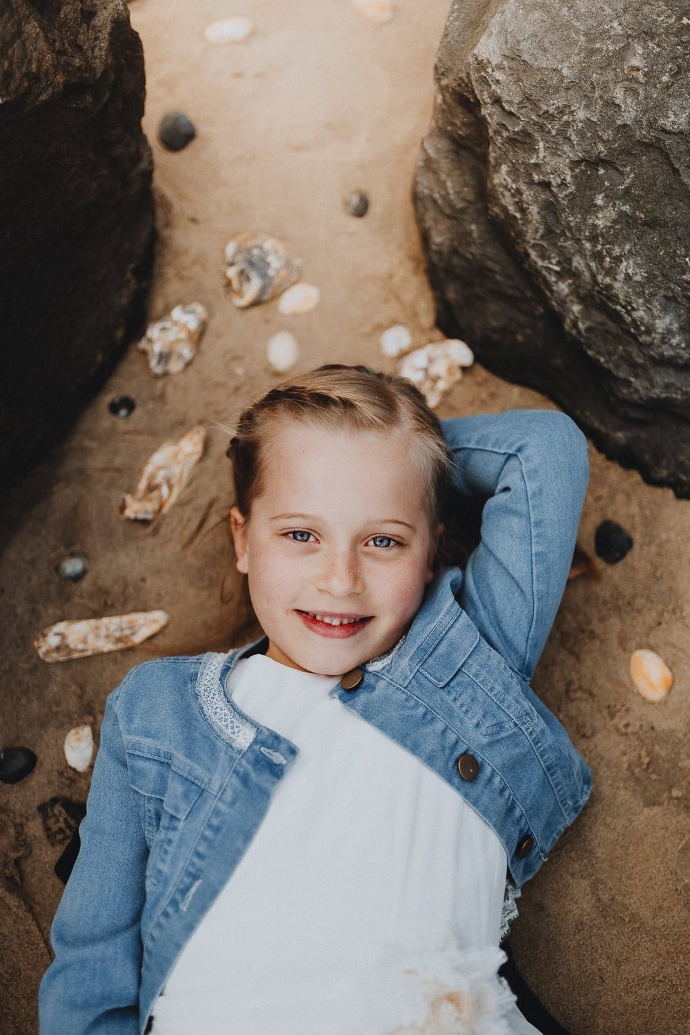 Meisje met blauwe ogen ligt ontspannen op het strand tussen schelpen, communie shoot in Koksijde, West-Vlaanderen