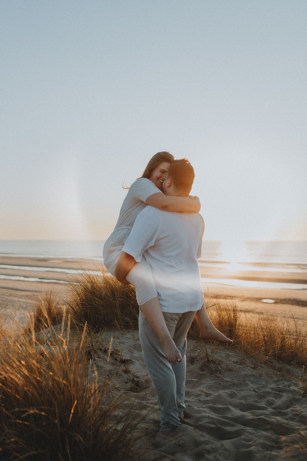 Koppel omhelst elkaar op het strand bij zonsondergang, de vrouw wordt opgetild, warme en romantische shoot in Koksijde, West-Vlaanderen.