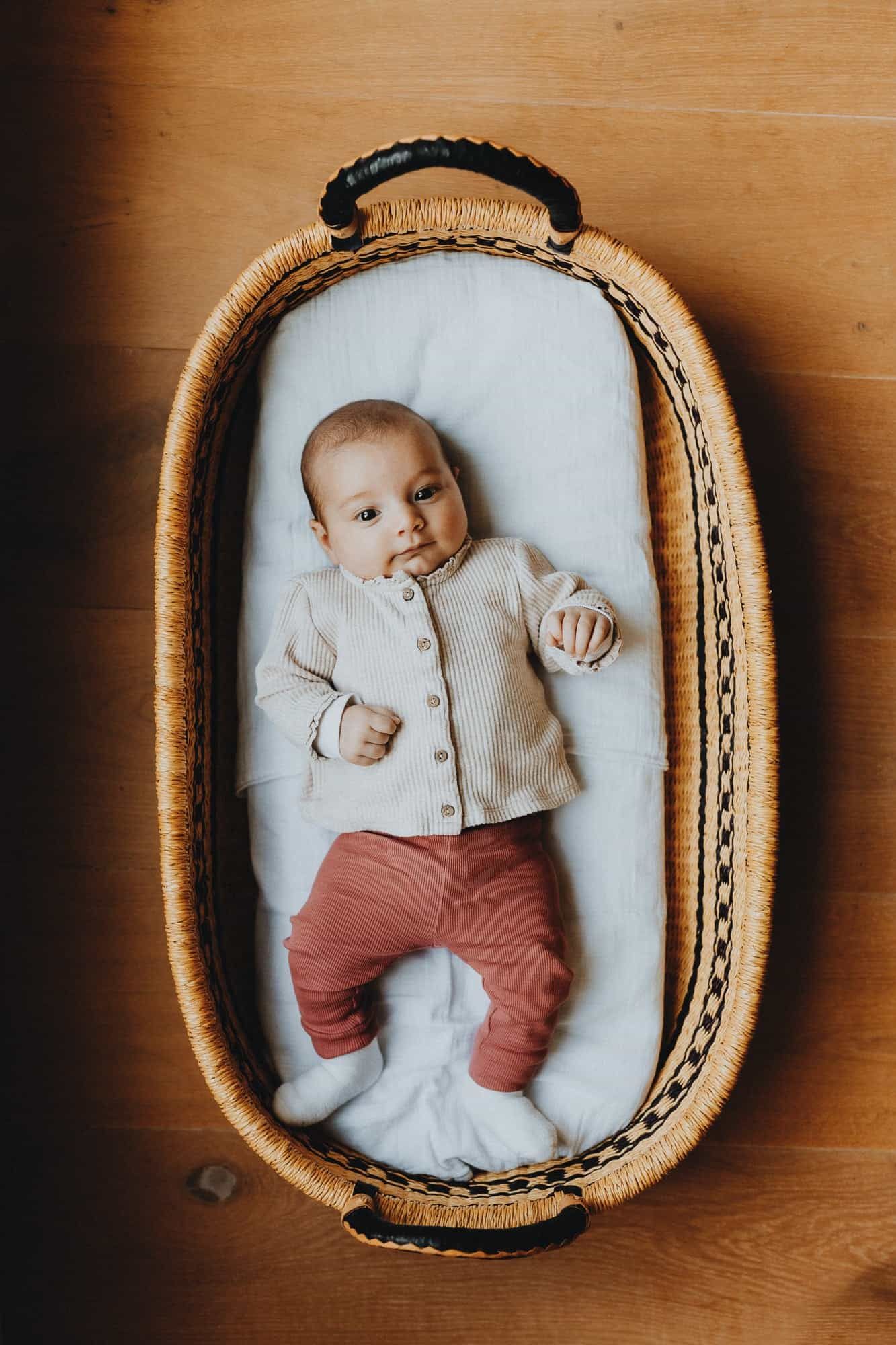 Newbornshoot in Kortemark: baby in een mand op houten vloer, met beige trui en rode broek. Kijkt rustig omhoog. Warme sfeer.