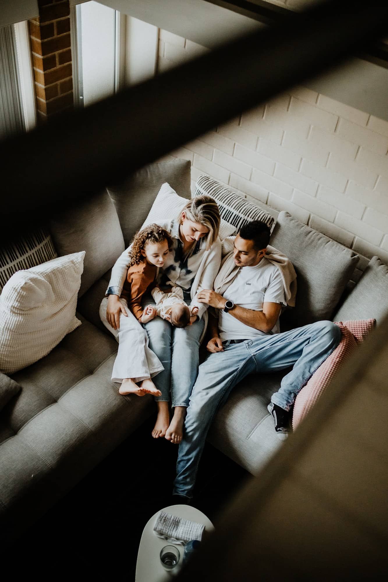 Een gezin van vier zit dicht bij elkaar op een grijze sofa in een warm verlichte kamer. Ouders en kind kijken liefdevol naar de baby.