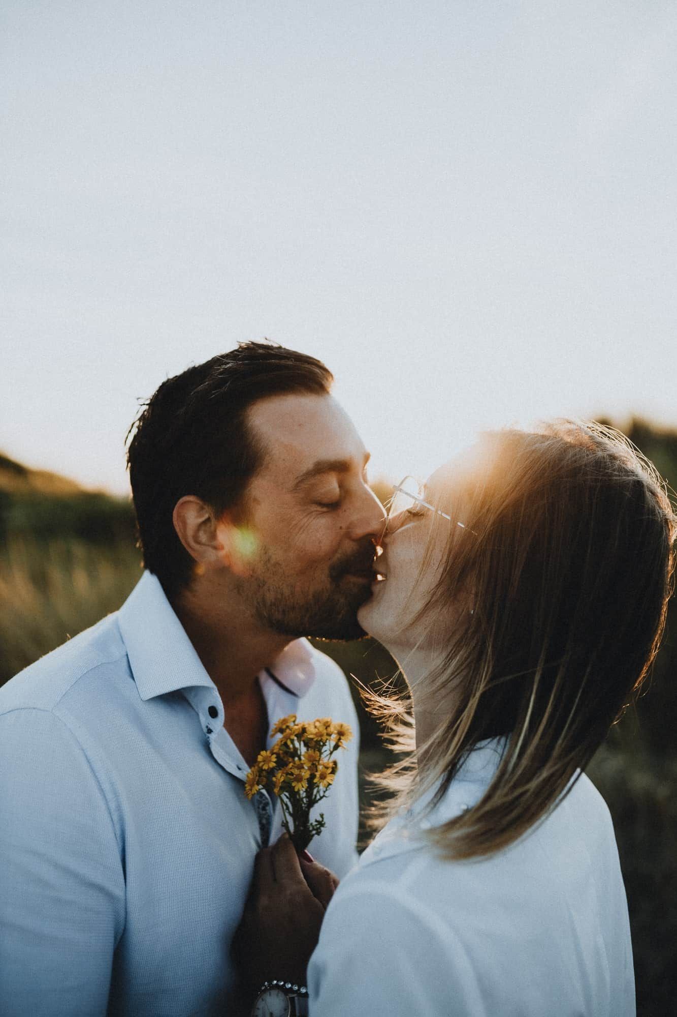 Een koppel kust teder in een zonovergoten veld, met gele bloemen in de hand. Warm licht creëert een romantische en intieme sfeer.