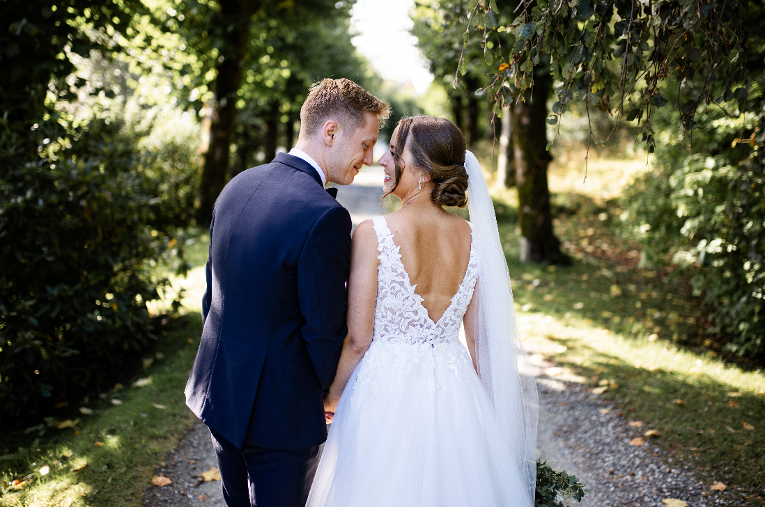 Wedding couple walking down a tree-lined path, bride in white gown with open back and groom in navy suit.