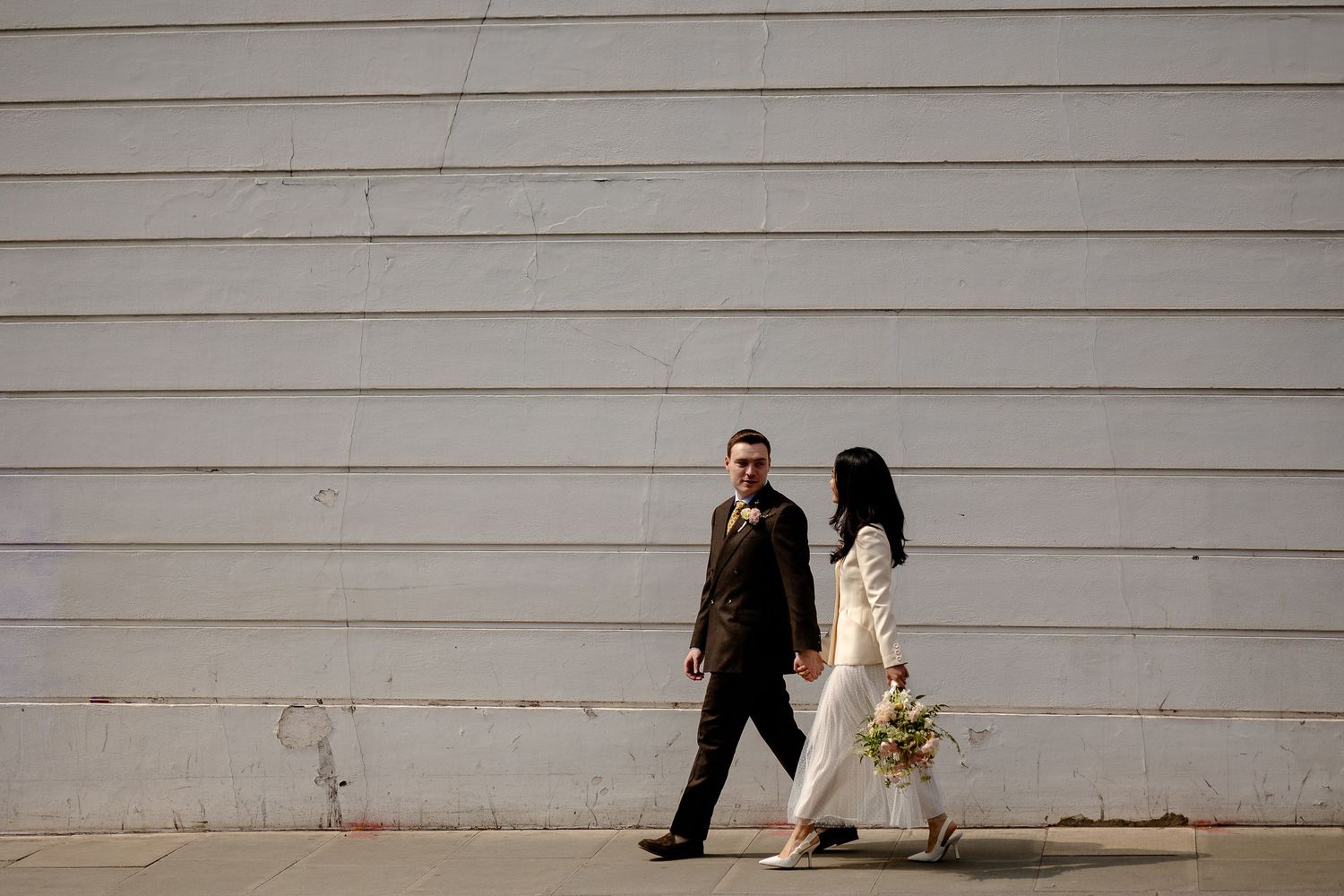 Two people in formal attire walk past white wall while one carries a bouquet of flowers.
