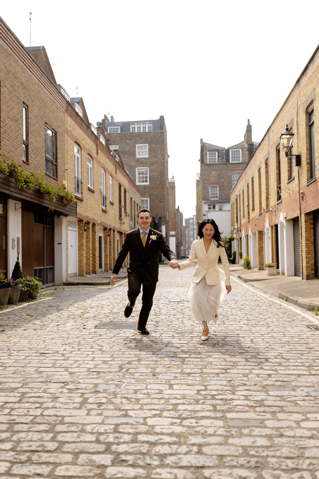 A couple runs playfully down a charming cobblestone street lined with historic brick buildings.