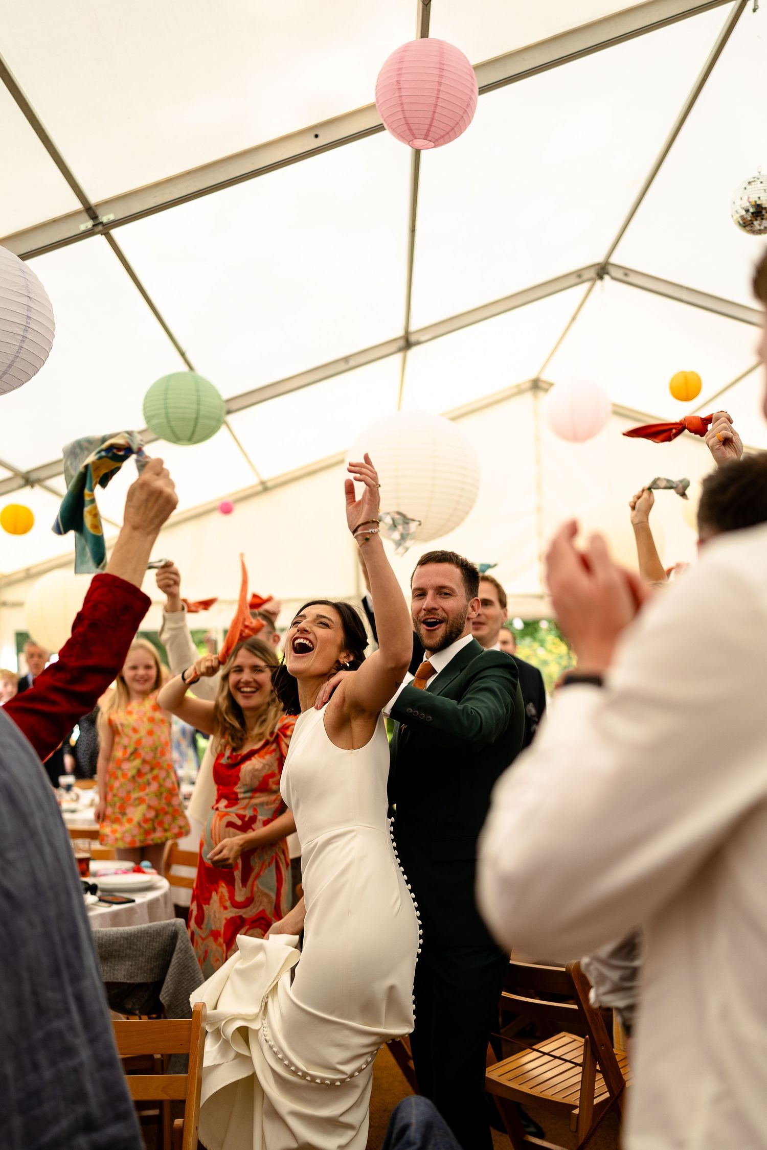 A crowd of wedding guests celebrate and dance joyfully under a tent with pink and white paper lanterns decorating the ceiling.