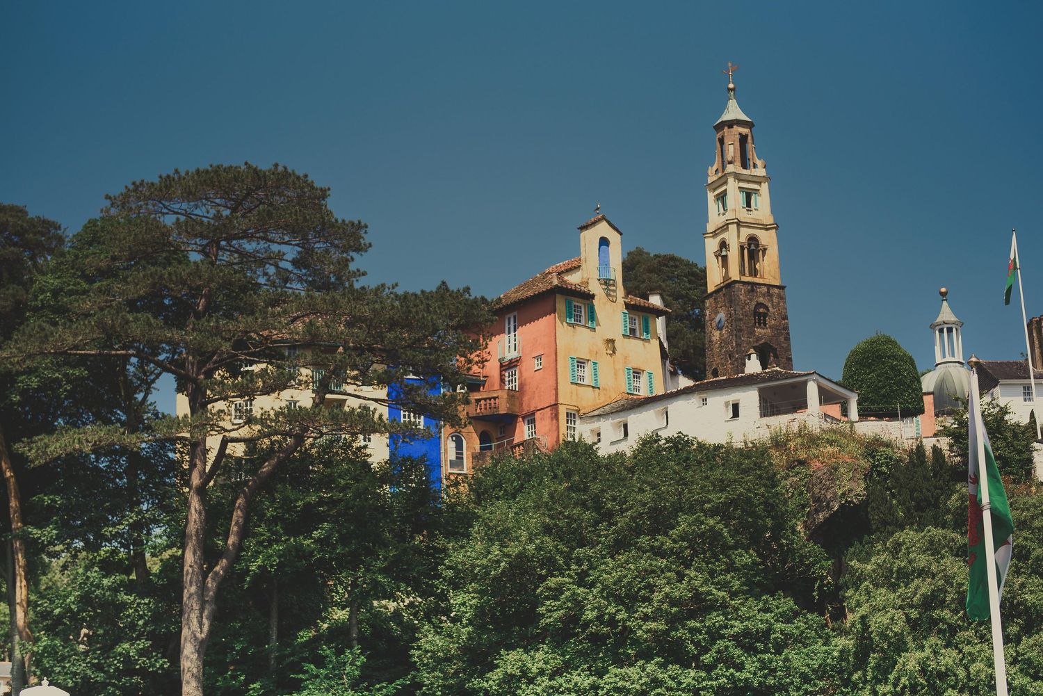 Historic church tower and colorful buildings perched on a lush hillside in Portmeirion, Wales against blue sky.