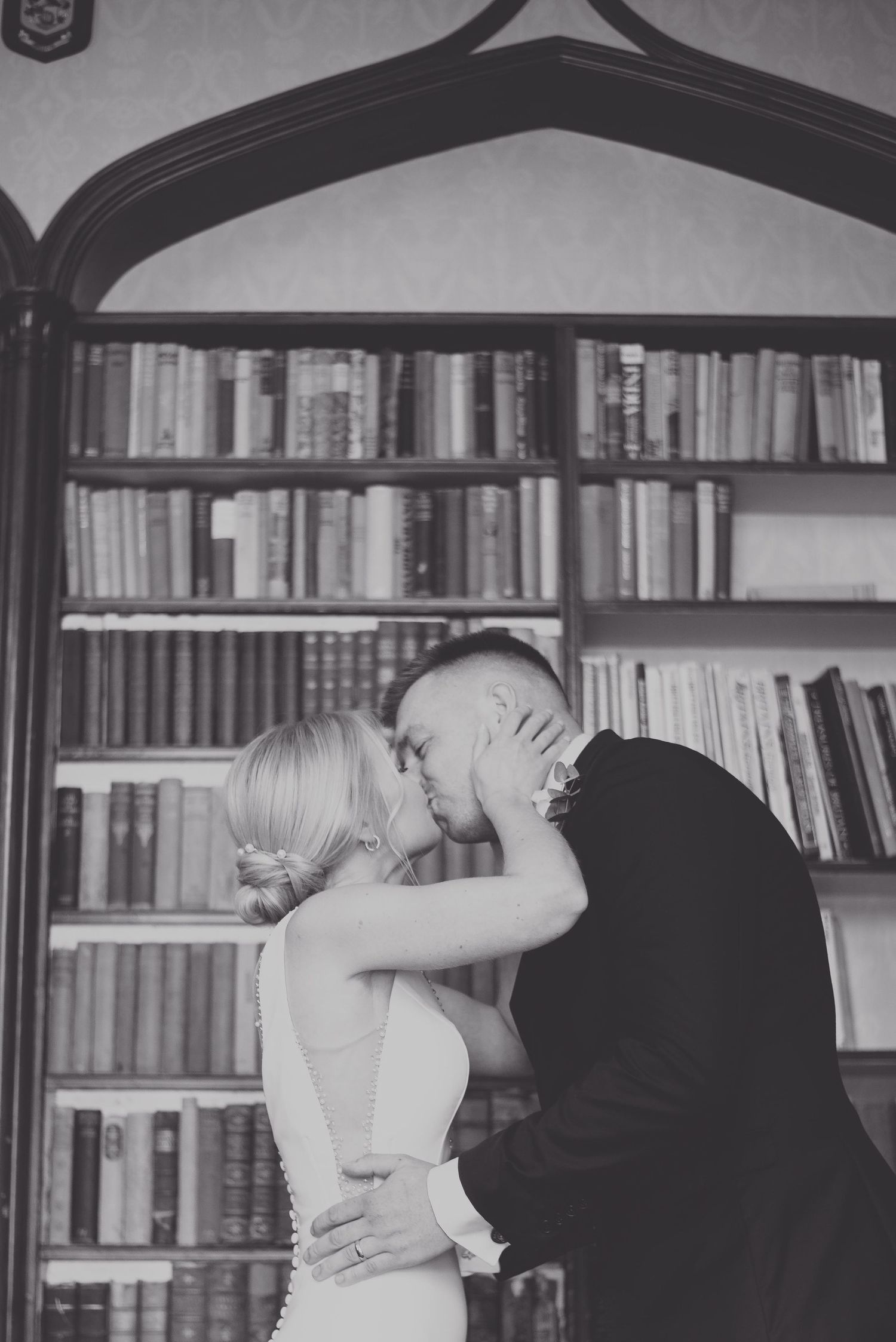 A romantic black and white photograph of a couple kissing in front of a large wooden bookshelf filled with books.