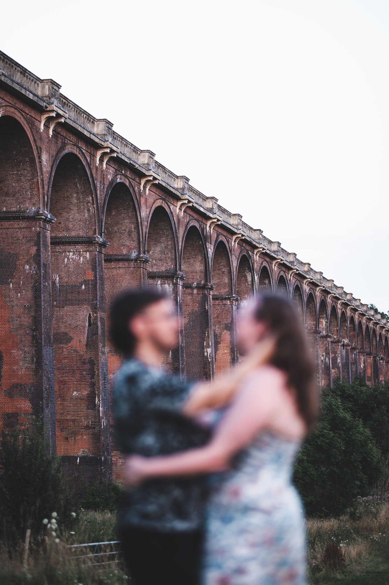 A couple embraces near historic brick railway viaduct arches at dusk.