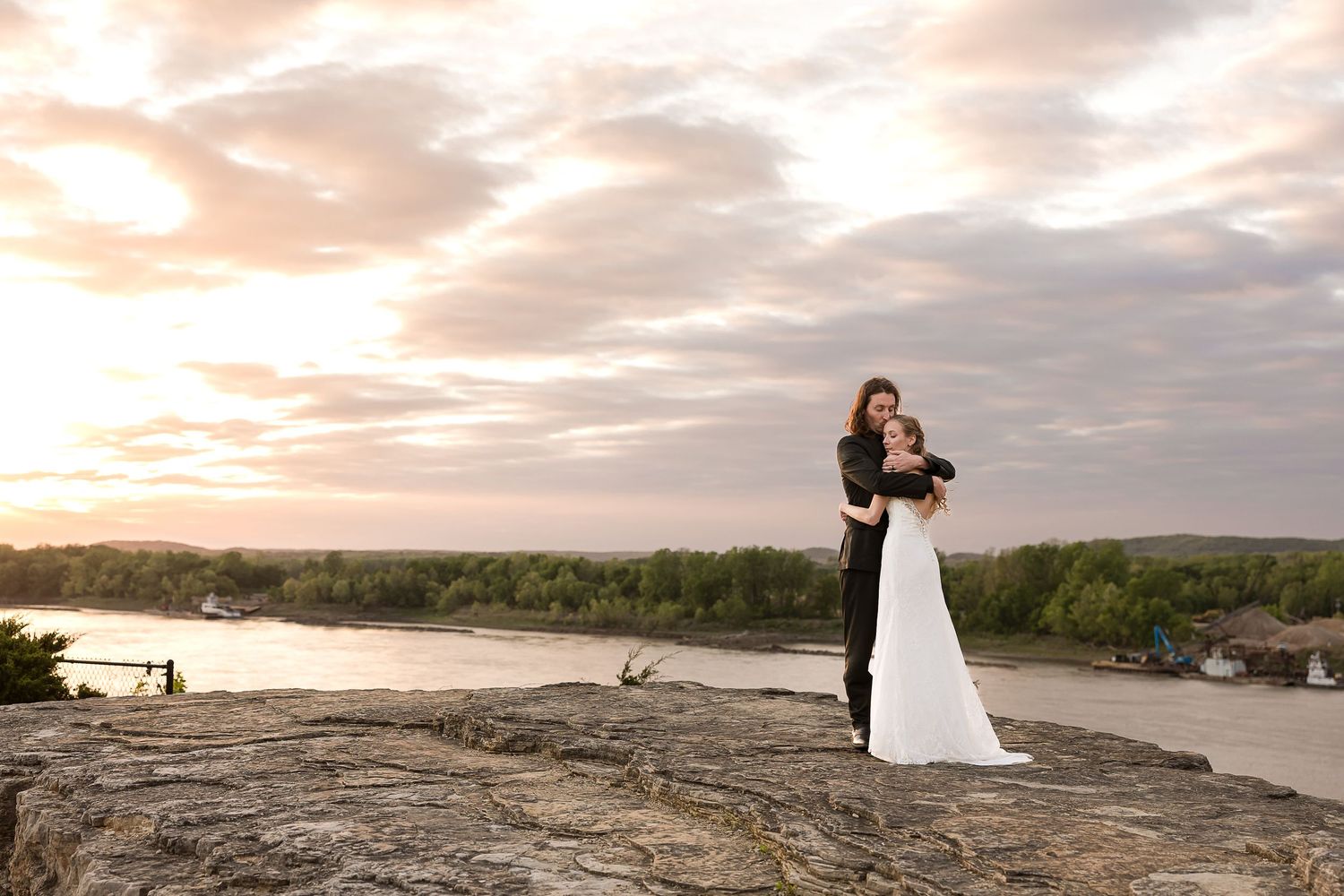 Silhouetted wedding couple stands on rocky overlook during dramatic sunset over river landscape.