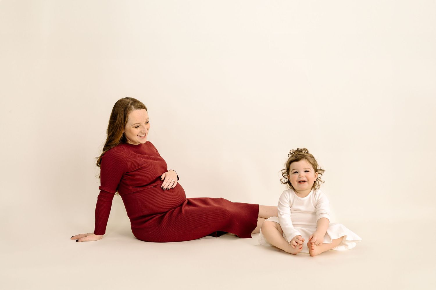 A child in white dress sits on cream floor while someone in burgundy dress reclines beside them against neutral backdrop.