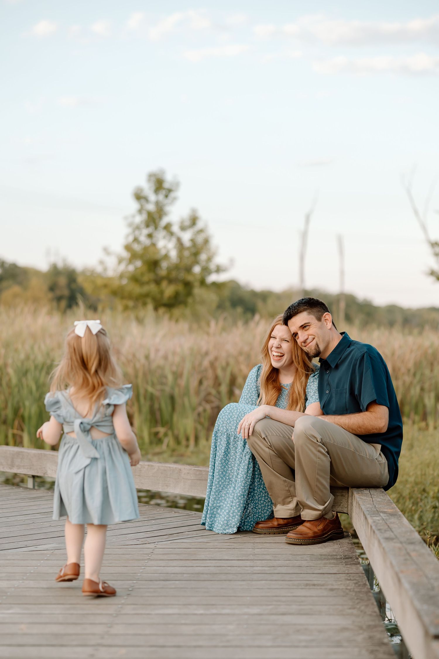 A scenic family moment captured on a wooden boardwalk surrounded by tall grasses at sunset.