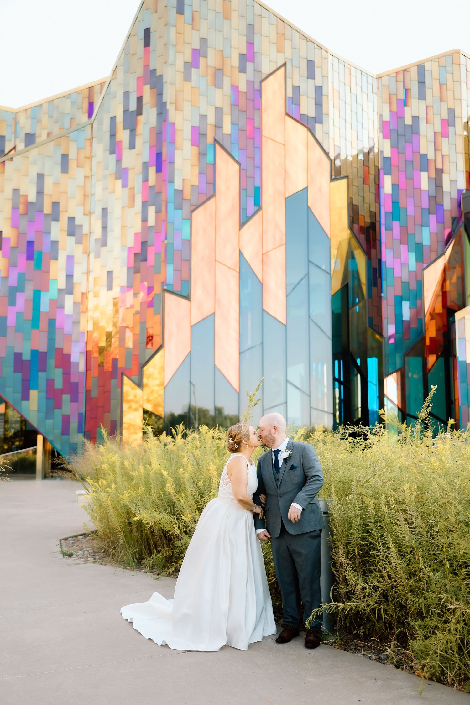 Wedding couple poses in front of a modern building with colorful geometric patterns and wild grasses.