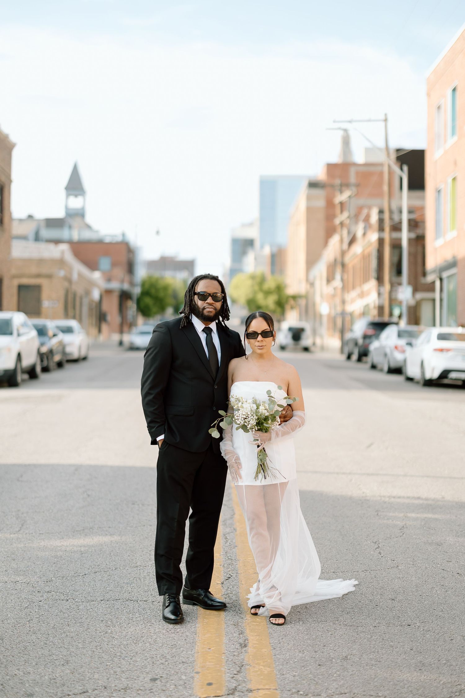 Couple poses in formal wedding attire on an urban street with parked cars and city buildings in the background.