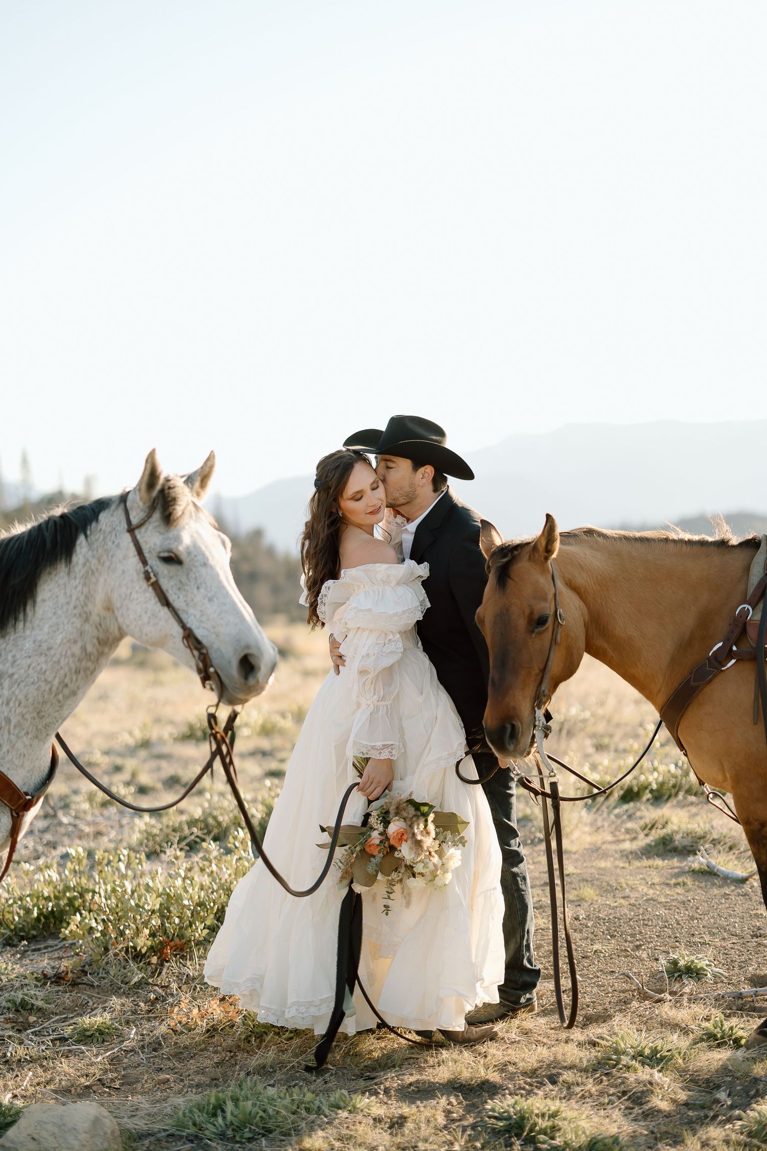 Western wedding couple with white and brown horses against mountain backdrop.