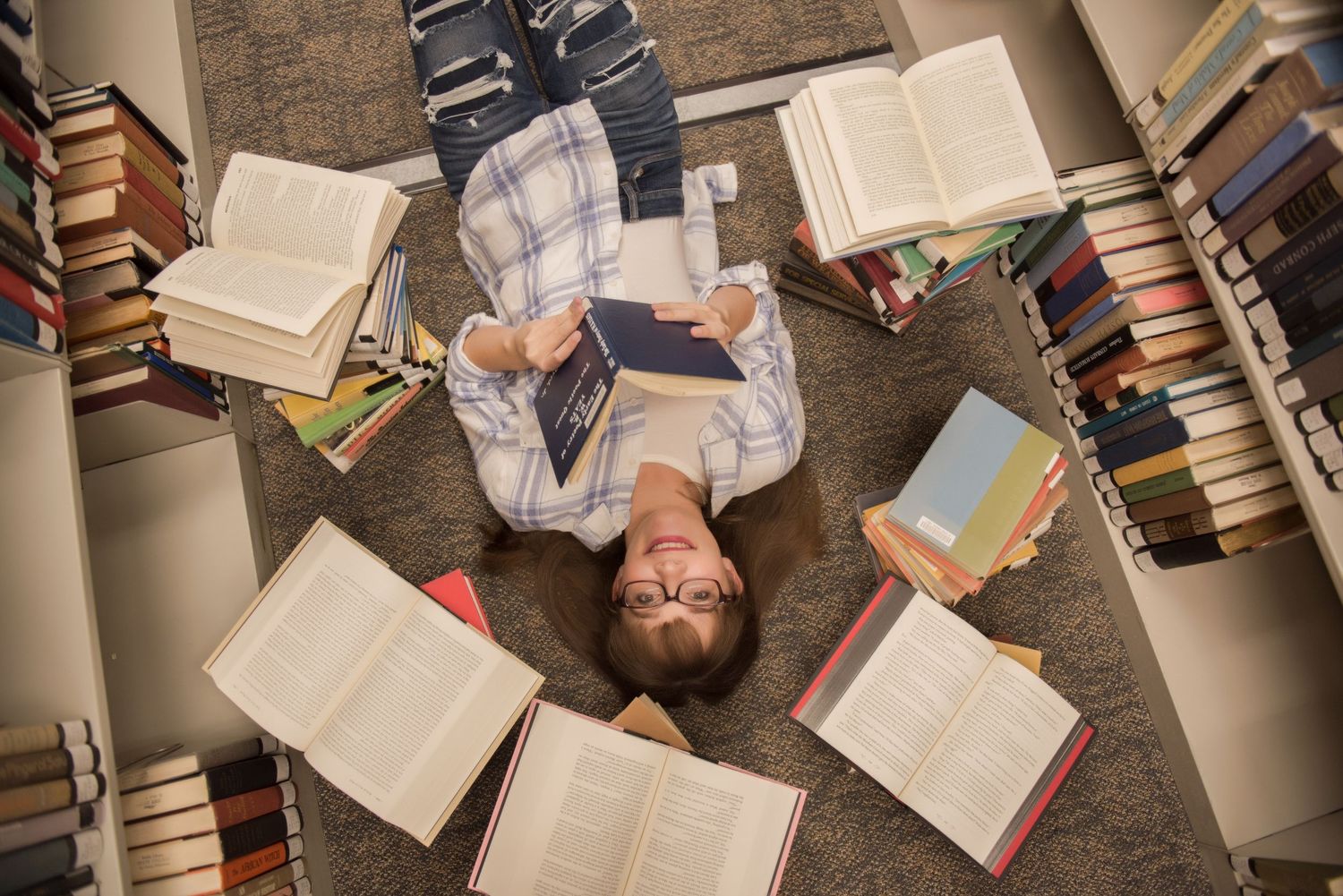A student lies on the floor surrounded by open books stacked in circular formation.