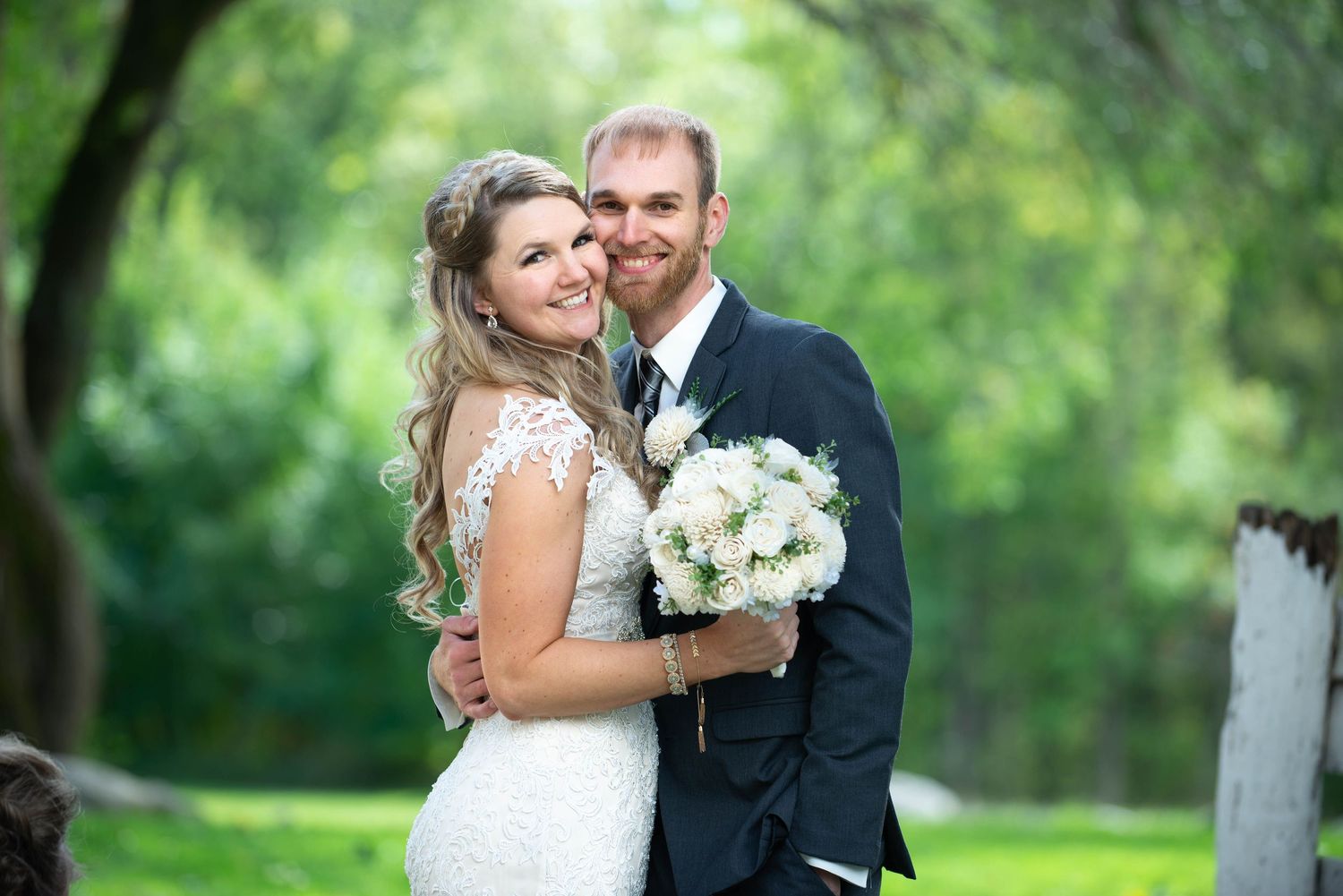 A happy wedding couple embraces while holding a white floral bouquet in a lush green garden. Rum River Barn Milaca Minnesota Weddings